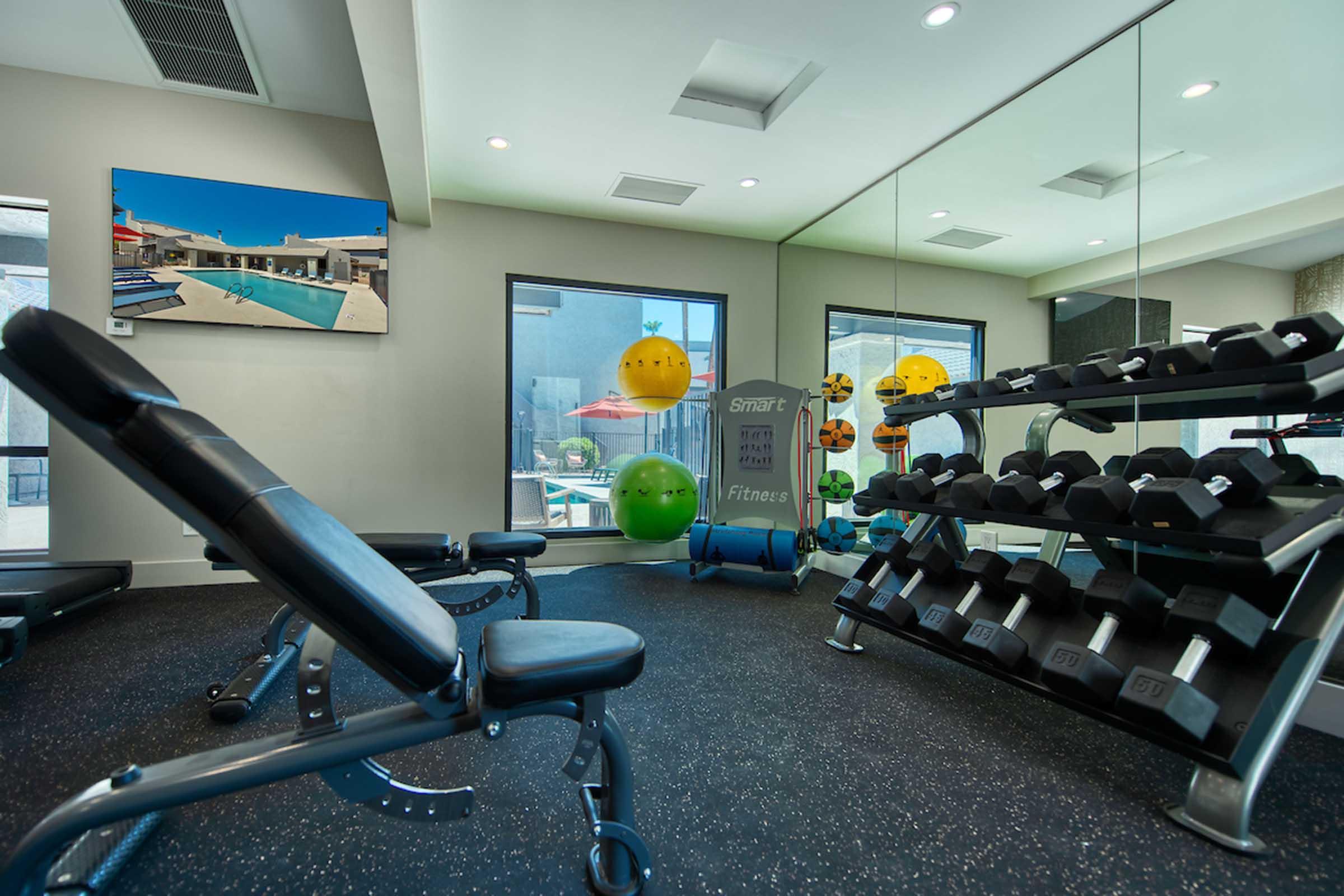 A modern gym space featuring a weight bench and a rack of dumbbells. The room has mirrors, colorful exercise balls, and motivational fitness posters. Natural light enters through large windows, creating an inviting atmosphere.