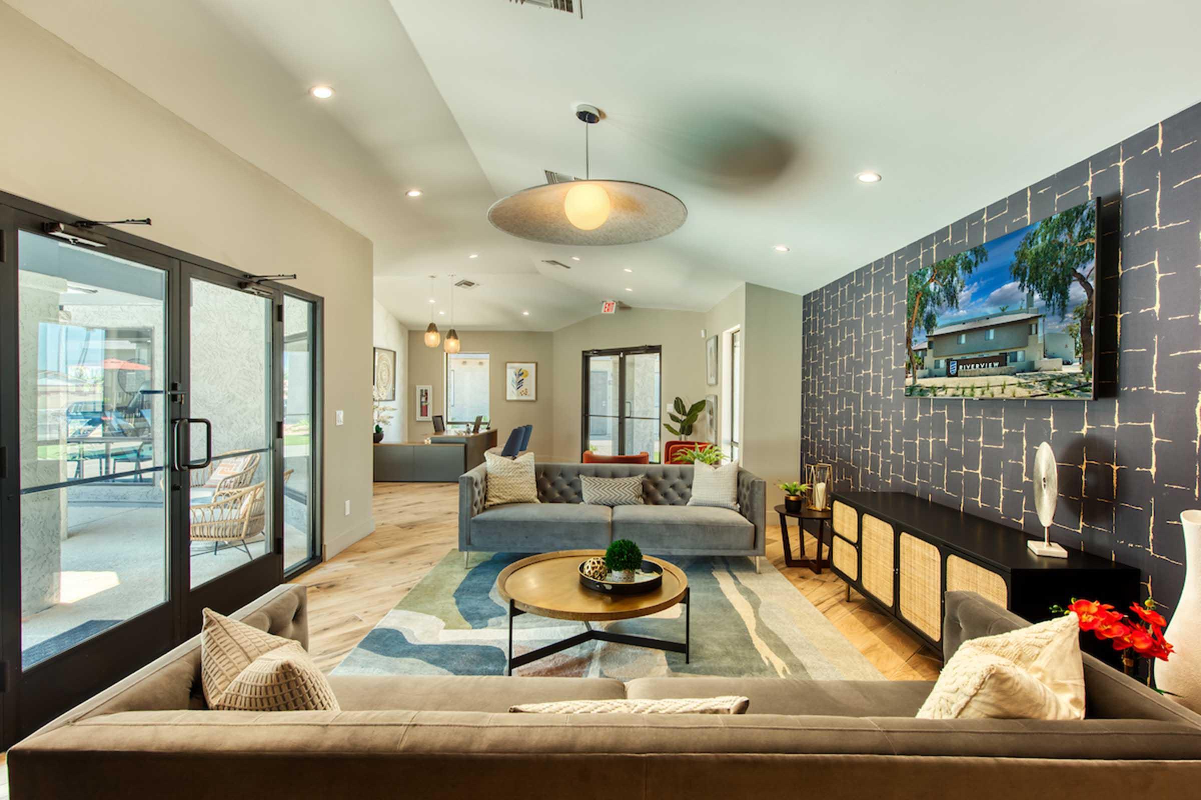 Modern lounge area with two sofas, a circular coffee table, and decorative pillows. A large wall-mounted TV displays a landscape, while a textured accent wall adds visual interest. Natural light fills the space through glass doors leading to an outdoor area, enhancing the inviting atmosphere.