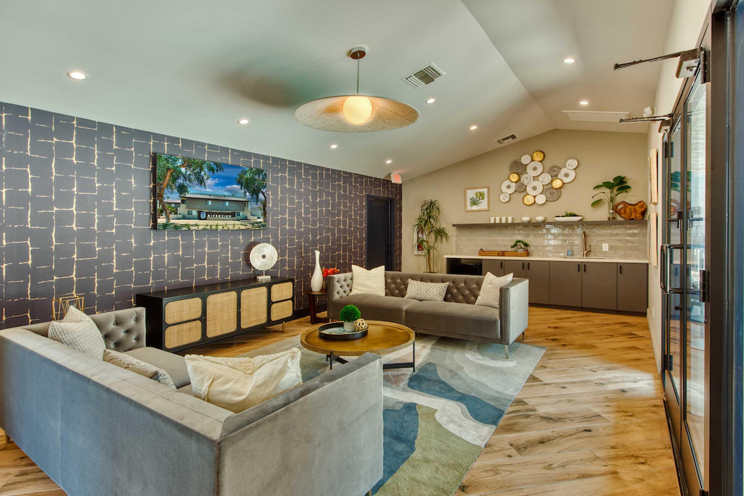 A modern living space featuring light-colored sofas, a round coffee table, and decorative wall art. The room has wood flooring, a ceiling light fixture, and a large TV mounted on the wall. There is a small kitchenette area with plants and decorative items in the background.
