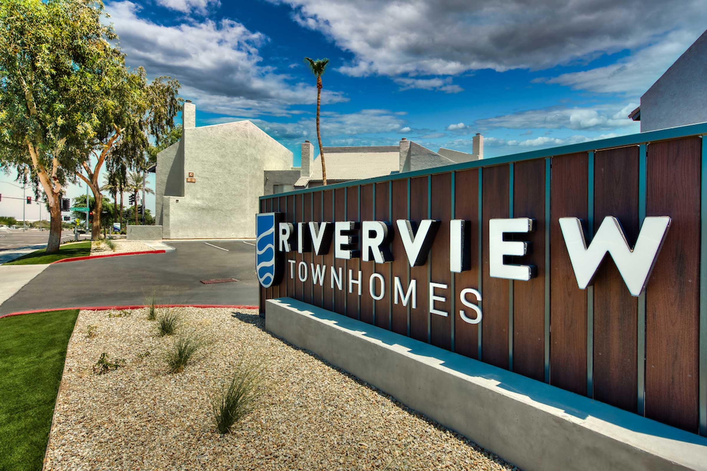 Sign for Riverview Townhomes with modern lettering, surrounded by landscaping and palm trees. The background features cloudy blue skies and residential buildings.