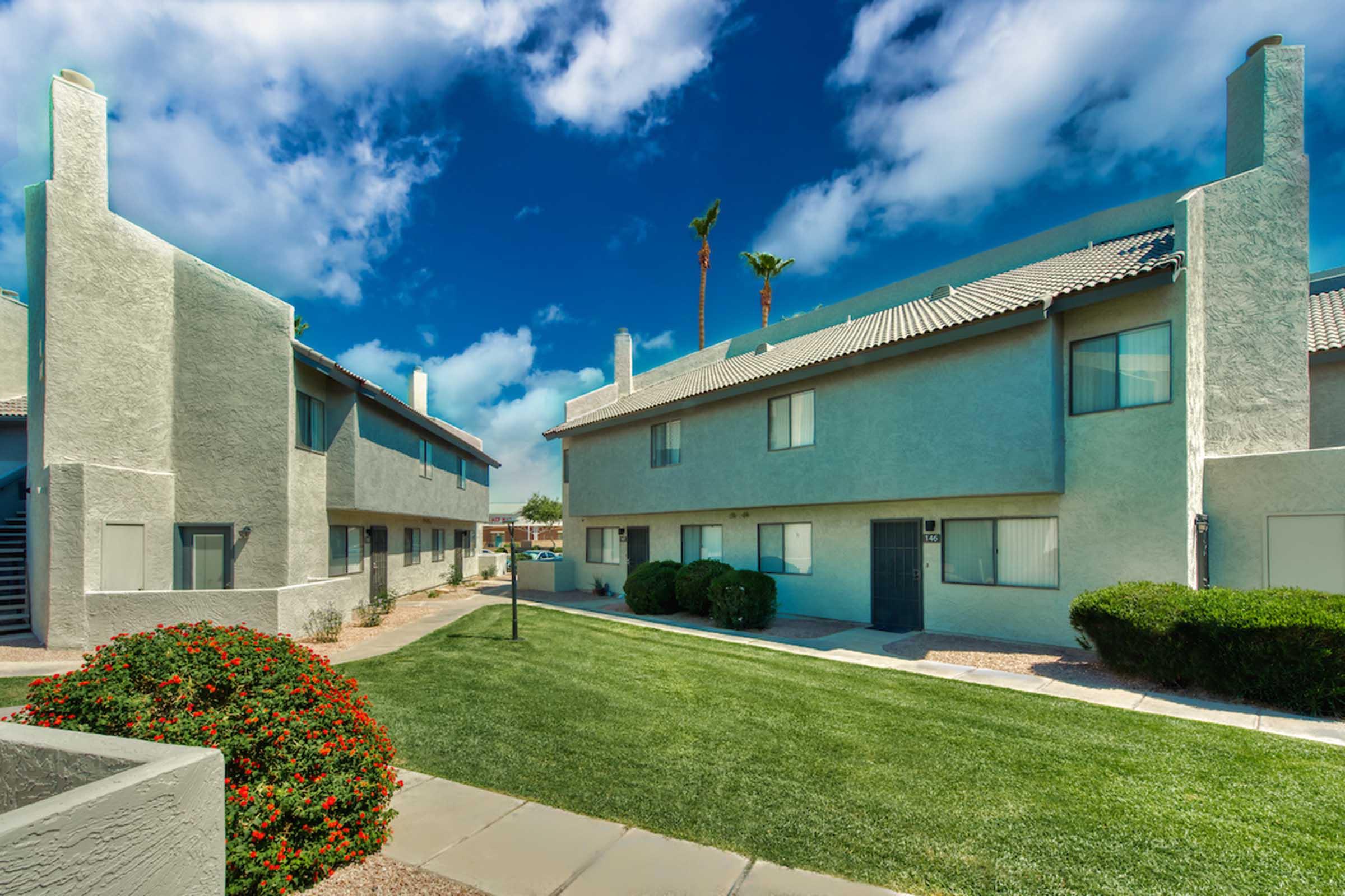 Apartment complex exterior featuring two-story buildings with gray walls and a well-maintained green lawn. The sky is bright with scattered clouds, and there are palm trees nearby. Flowering bushes add color to the landscape, creating a pleasant outdoor environment.
