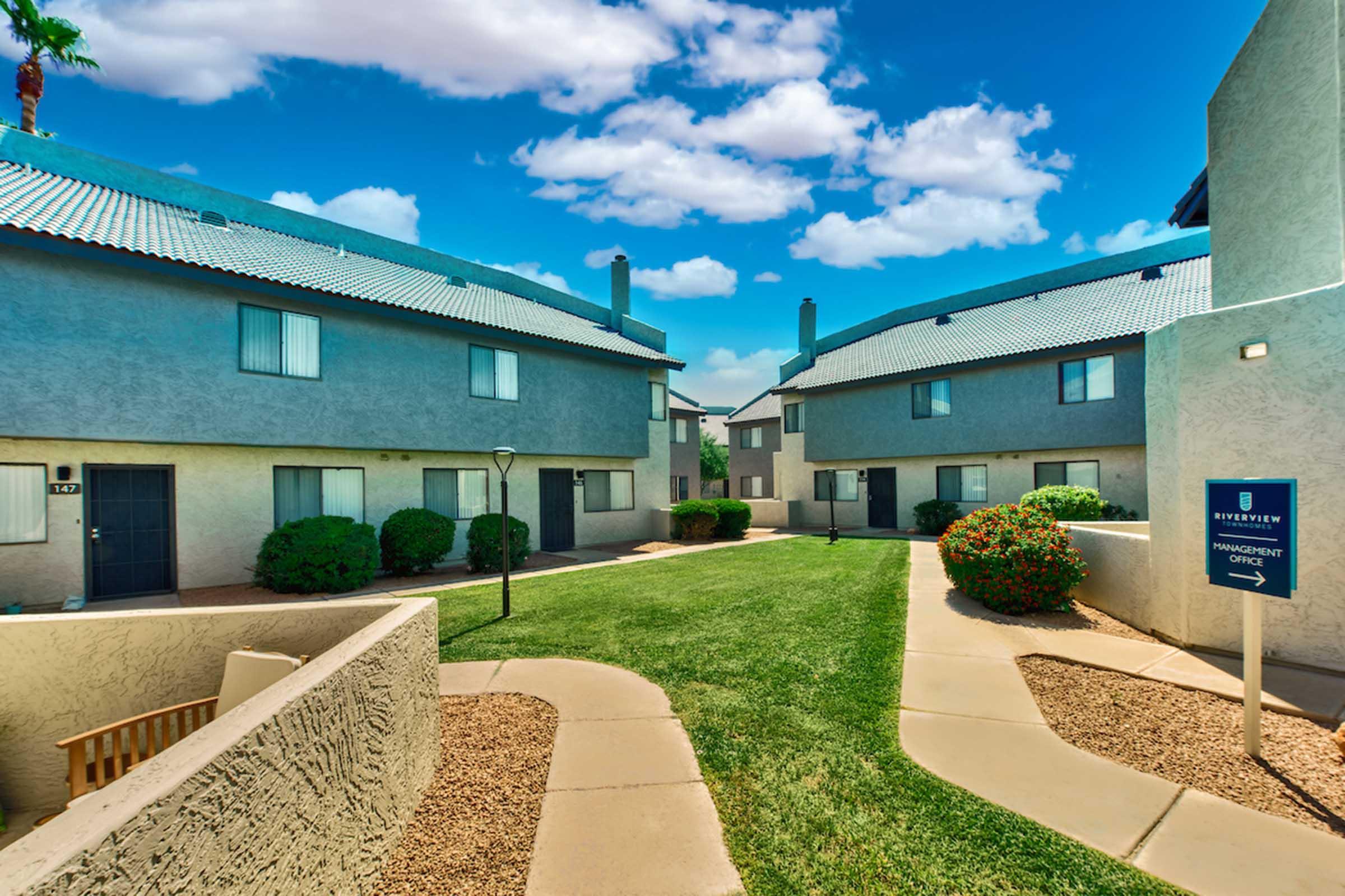 A view of a sunny courtyard featuring two-story apartment buildings in light colors, surrounded by well-maintained grass and shrubs. The sky is bright blue with fluffy clouds. A pathway winds through the landscape, leading to a sign for "Riverstein Improvement."