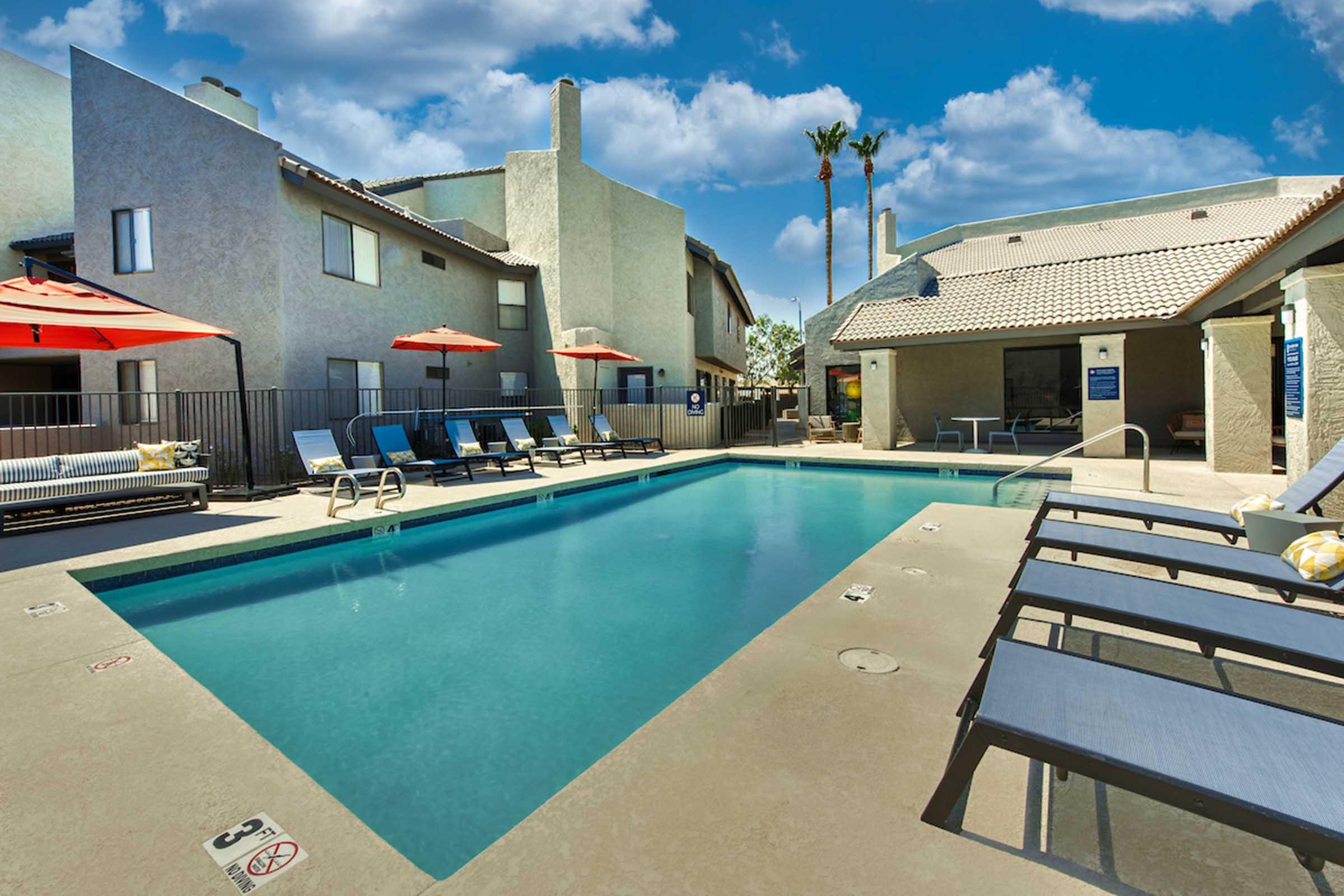 A swimming pool area with lounge chairs and orange umbrellas, surrounded by modern buildings. The scene is bright and sunny, featuring palm trees and a clear blue sky, creating a relaxing atmosphere. There are safety signs visible near the pool edge.