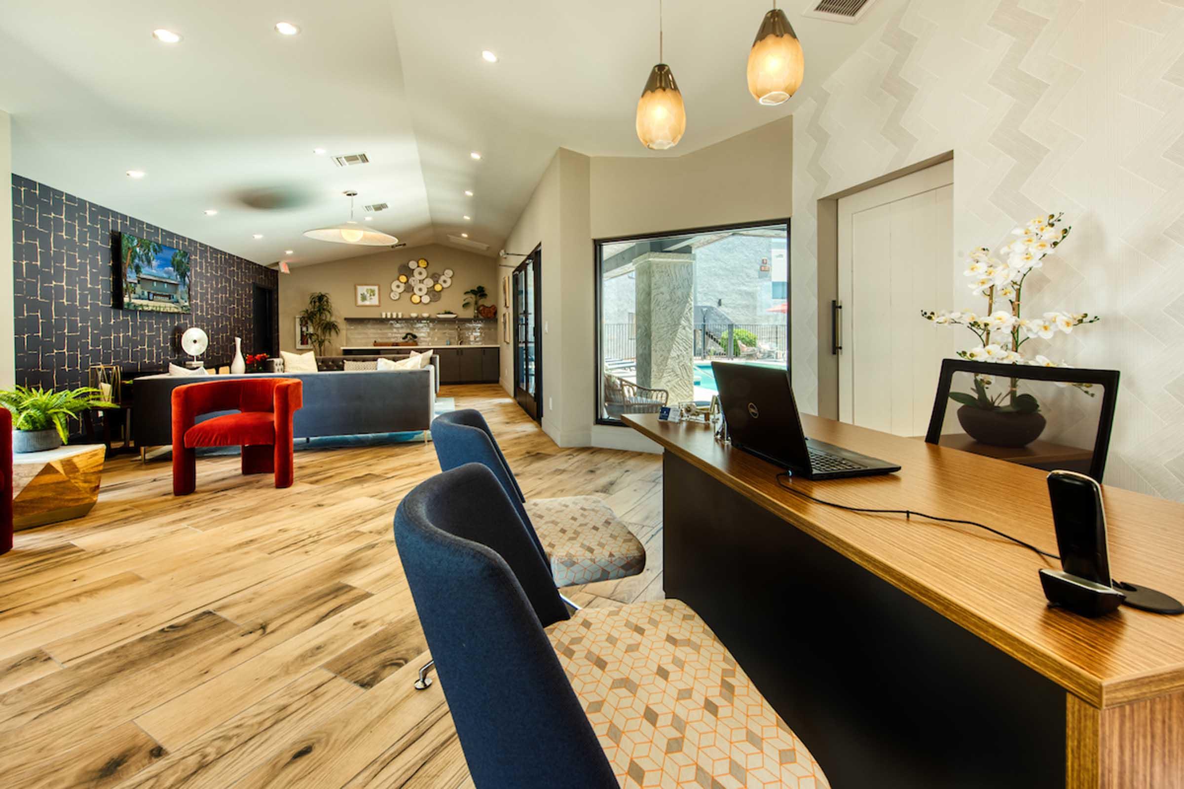 A modern reception area featuring a wooden desk with a laptop and phone. There are stylish chairs in blue and red, decorative plants, and wall art. Large windows allow natural light to fill the space, creating a welcoming atmosphere with wooden flooring.