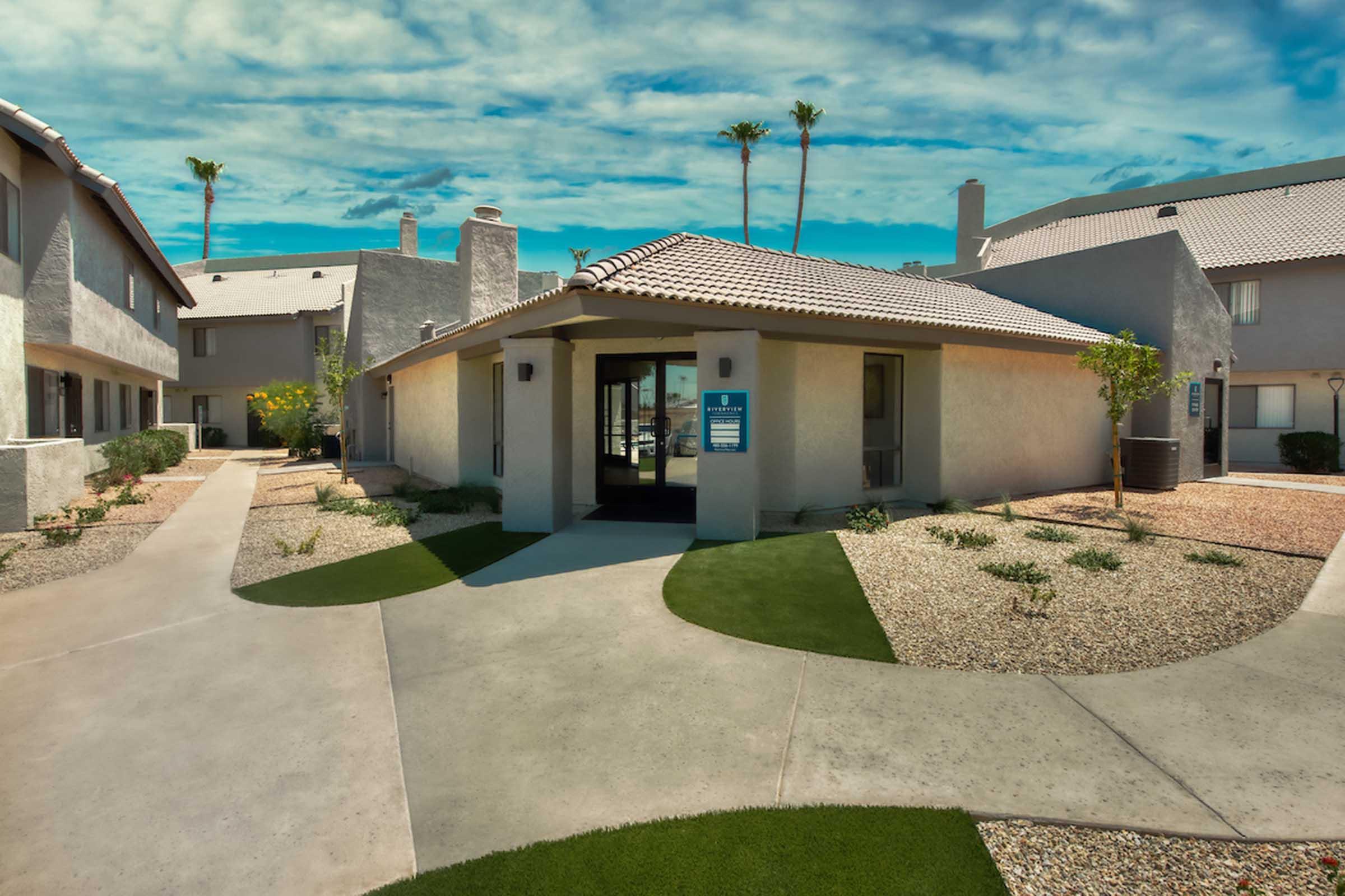 A modern, single-story building with a tile roof and large glass entrance. The surrounding area features landscaped pathways, green lawn patches, and decorative rocks. Palm trees are visible against a bright blue sky with fluffy clouds. The scene reflects a well-maintained residential complex.