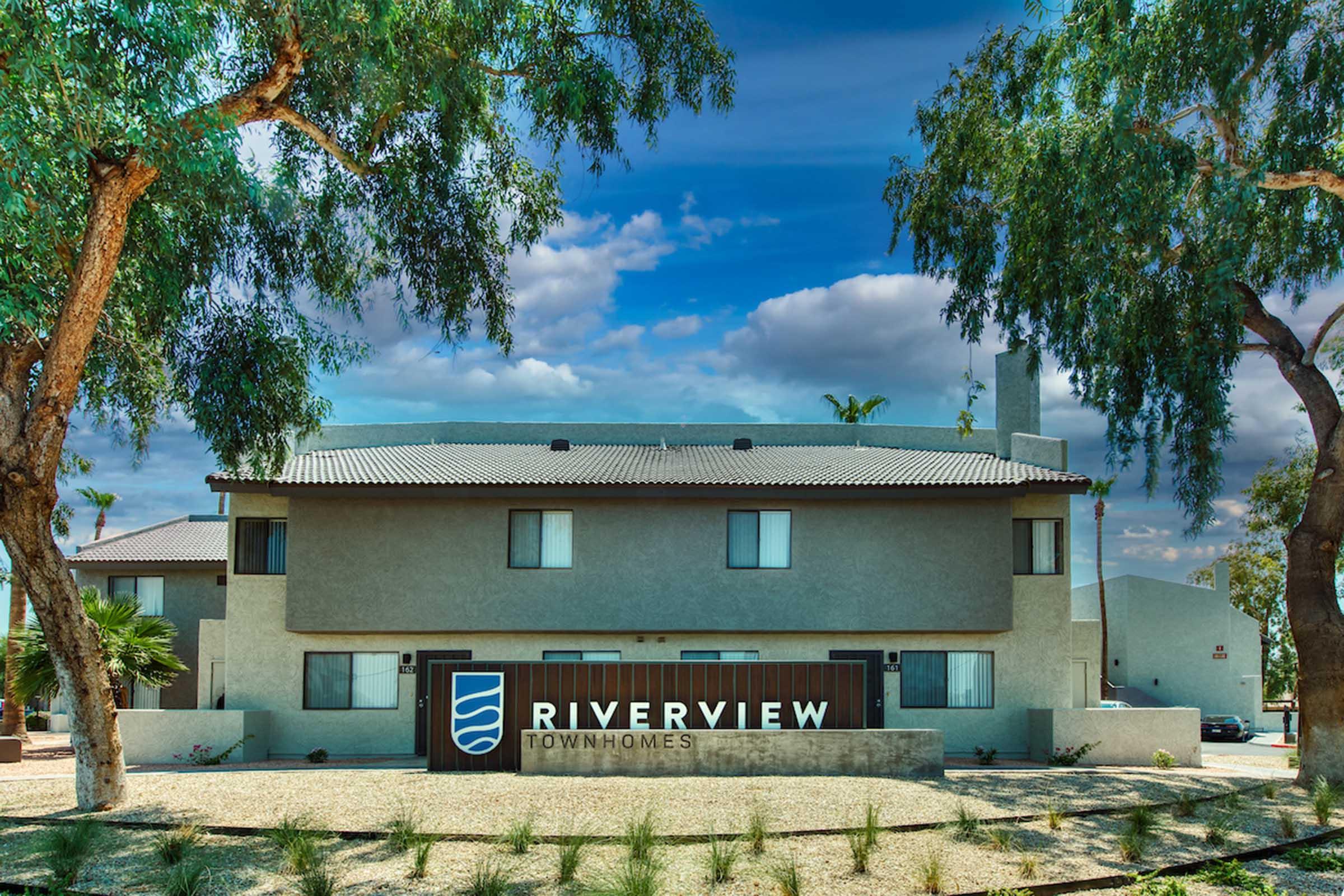 A modern residential building featuring the sign "RIVERVIEW TOWNHOMES." The building has multiple windows and a light exterior, surrounded by greenery and trees. The sky is partly cloudy with blue hues, creating a welcoming atmosphere for potential residents.