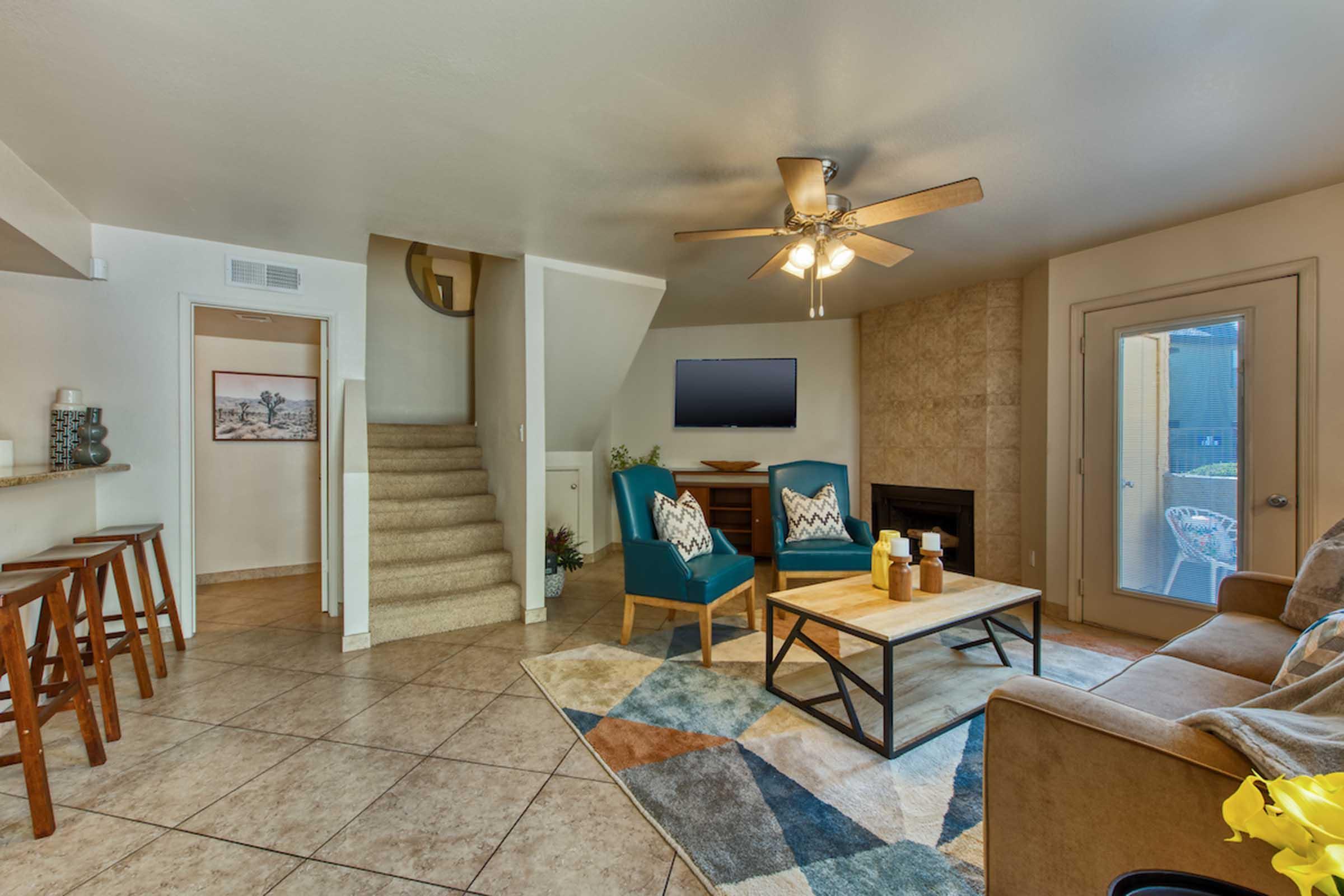 A cozy living room featuring a sofa, two blue accent chairs, and a coffee table with decorative items. A flat-screen TV is mounted on the wall above a stone fireplace. Light-colored stairs lead to an upper level, and a patio door opens to the outside. The space is decorated with a colorful area rug.