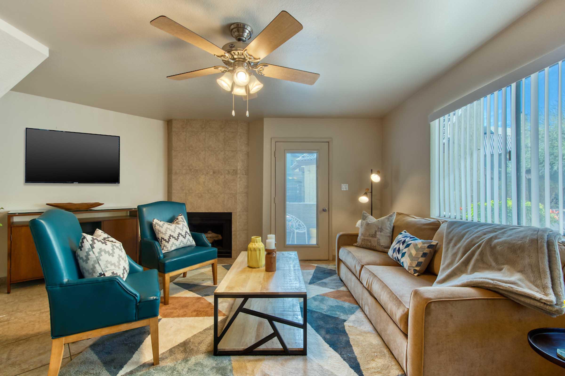 A modern living room featuring a beige sofa with decorative pillows, two teal armchairs, a geometric patterned rug, a coffee table with decorative vases, and a flat-screen TV mounted on the wall. Large windows allow natural light to fill the space, creating a warm and inviting atmosphere.