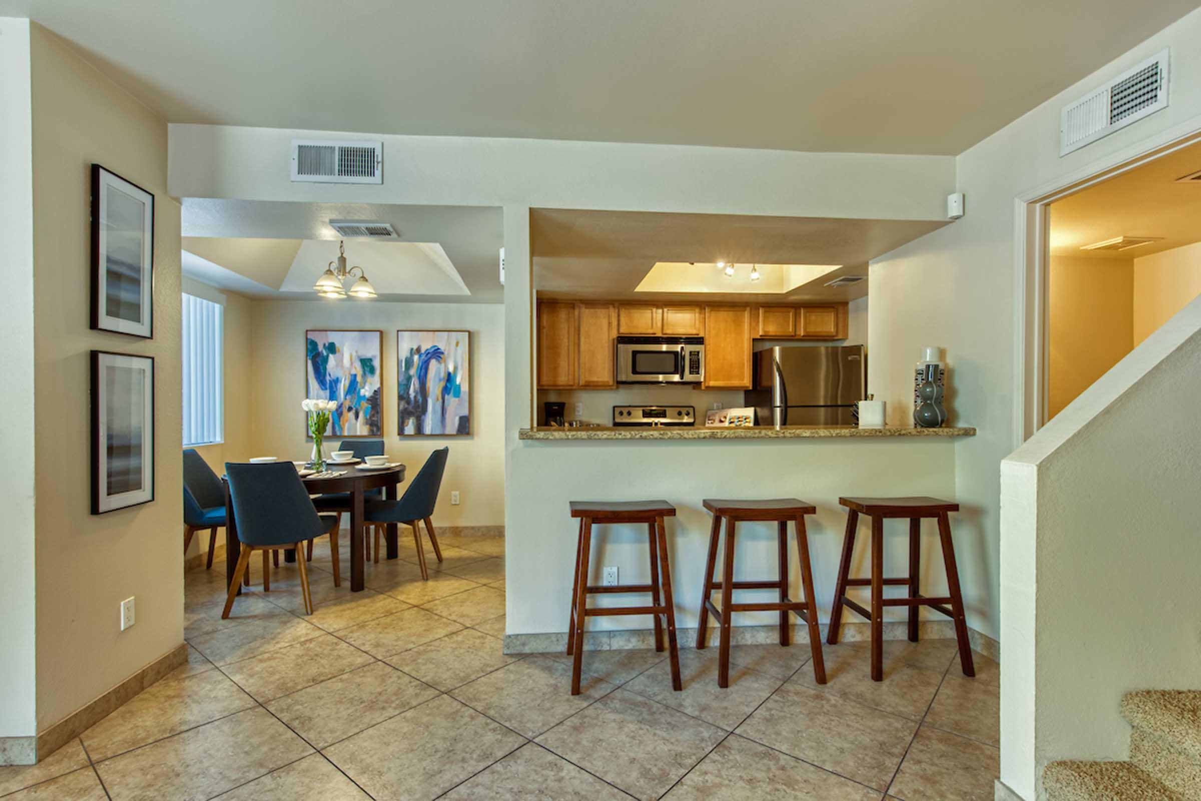 Interior of a modern kitchen and dining area. The kitchen features wooden cabinets, a stainless steel refrigerator, and a countertop with seating. A round dining table with blue chairs is set nearby, and colorful abstract artwork adorns the walls, enhancing the inviting ambiance.