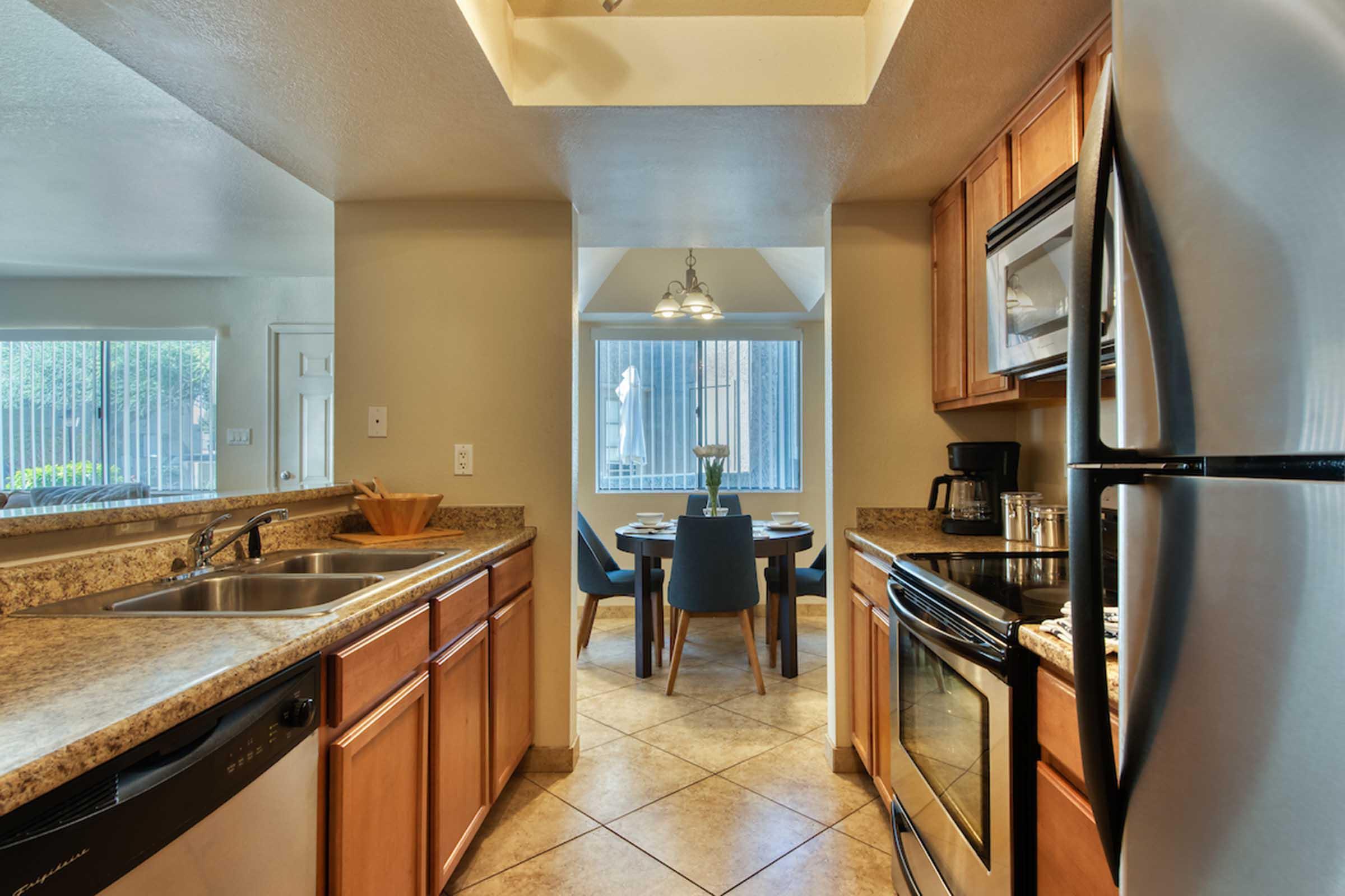 A modern kitchen with wooden cabinets, granite countertops, and stainless steel appliances. Natural light enters through windows, illuminating a dining area with a round table and chairs. The layout is open and inviting, featuring a sink and dishwasher on one side and a stove and microwave on the other.