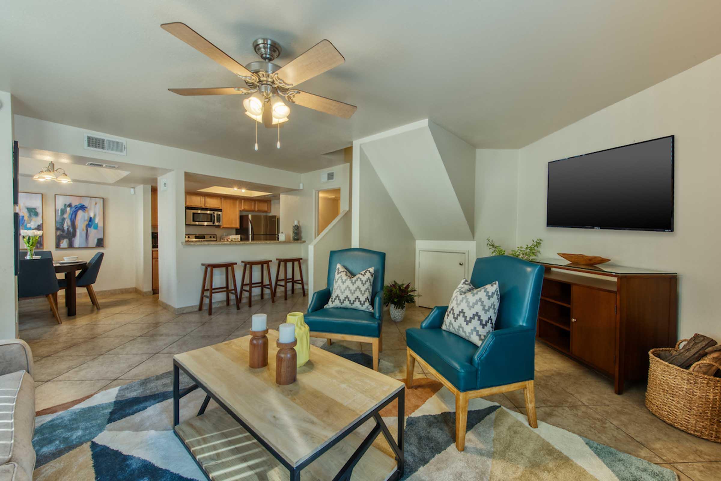 A modern living room featuring blue accent chairs, a wooden coffee table with decorative items, and a ceiling fan. The space has tile flooring, light-colored walls, and a flat-screen TV mounted on the wall. In the background, a dining area with a table and kitchen can be seen.