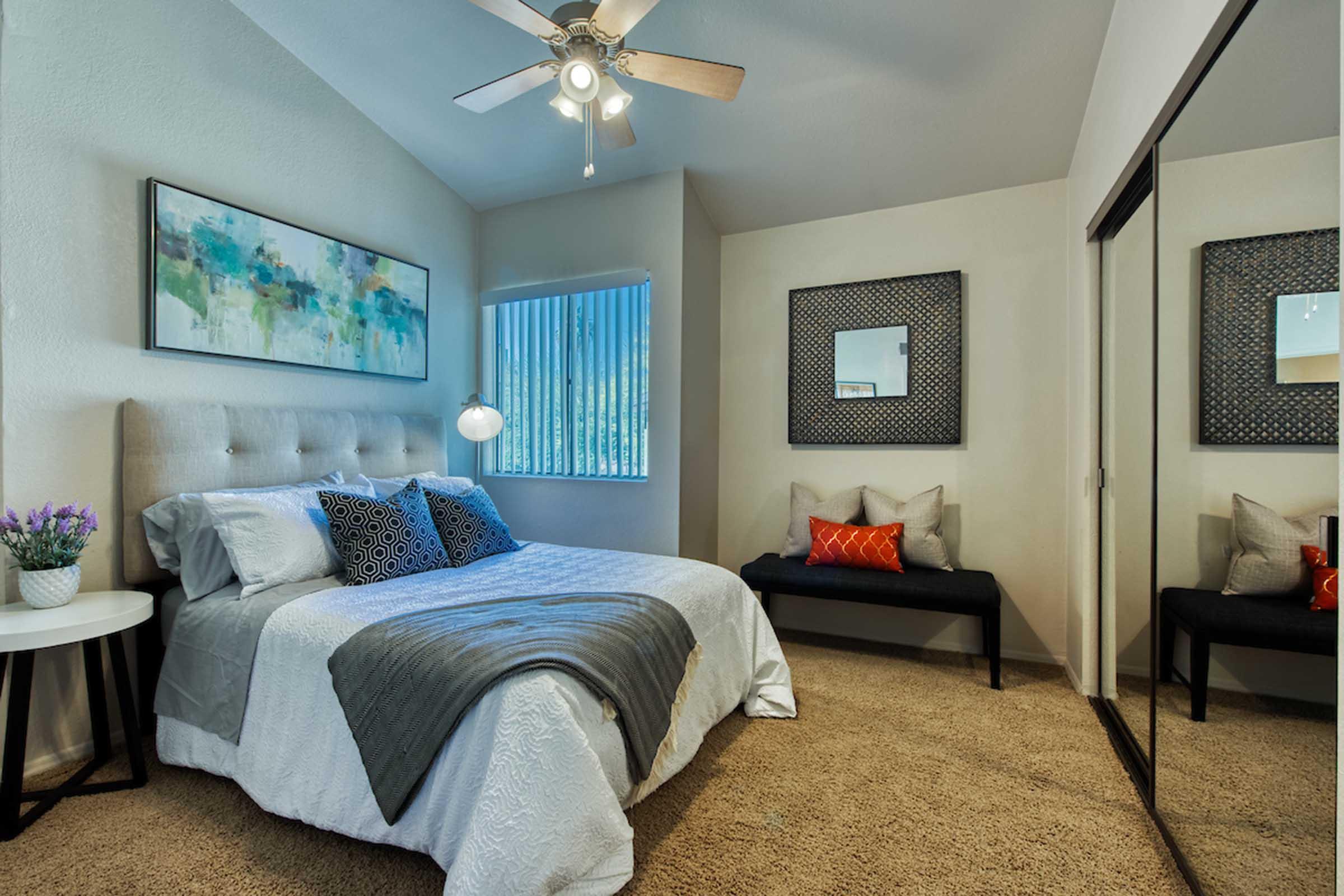 A cozy bedroom featuring a queen-sized bed with light gray bedding, decorative pillows, and a soft throw. There's a stylish bed frame, a nightstand with a lamp, and a large mirror on the closet doors. Two framed artworks adorn the walls, and a small bench with colorful cushions sits by the window, which has vertical blinds.