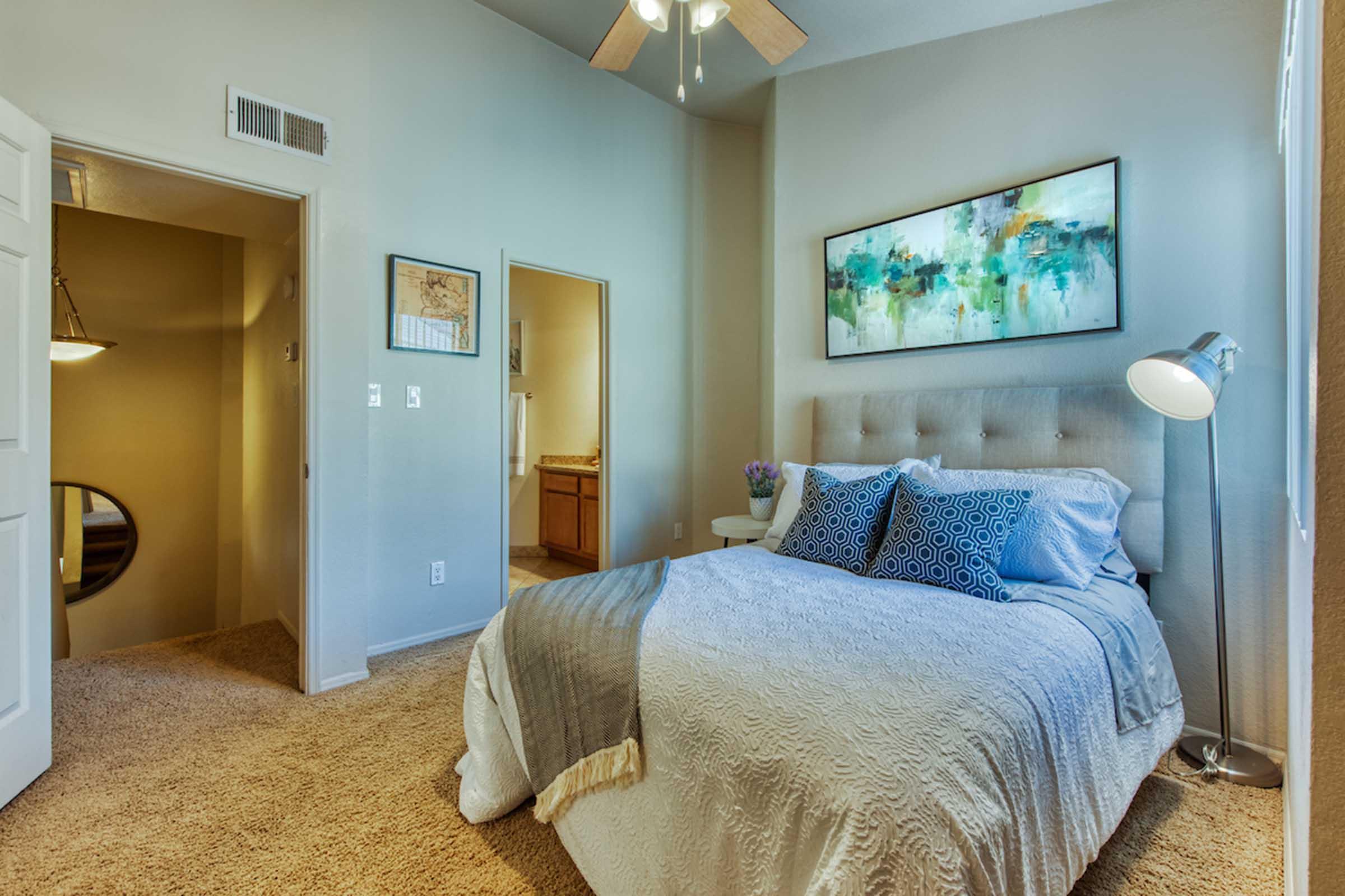 A cozy bedroom featuring a light-colored bed with decorative pillows, a bedside lamp, and a piece of abstract wall art. The room has warm, neutral walls and carpet, with a door leading to a bathroom and a glimpse of a staircase in the background. Soft lighting creates an inviting atmosphere.