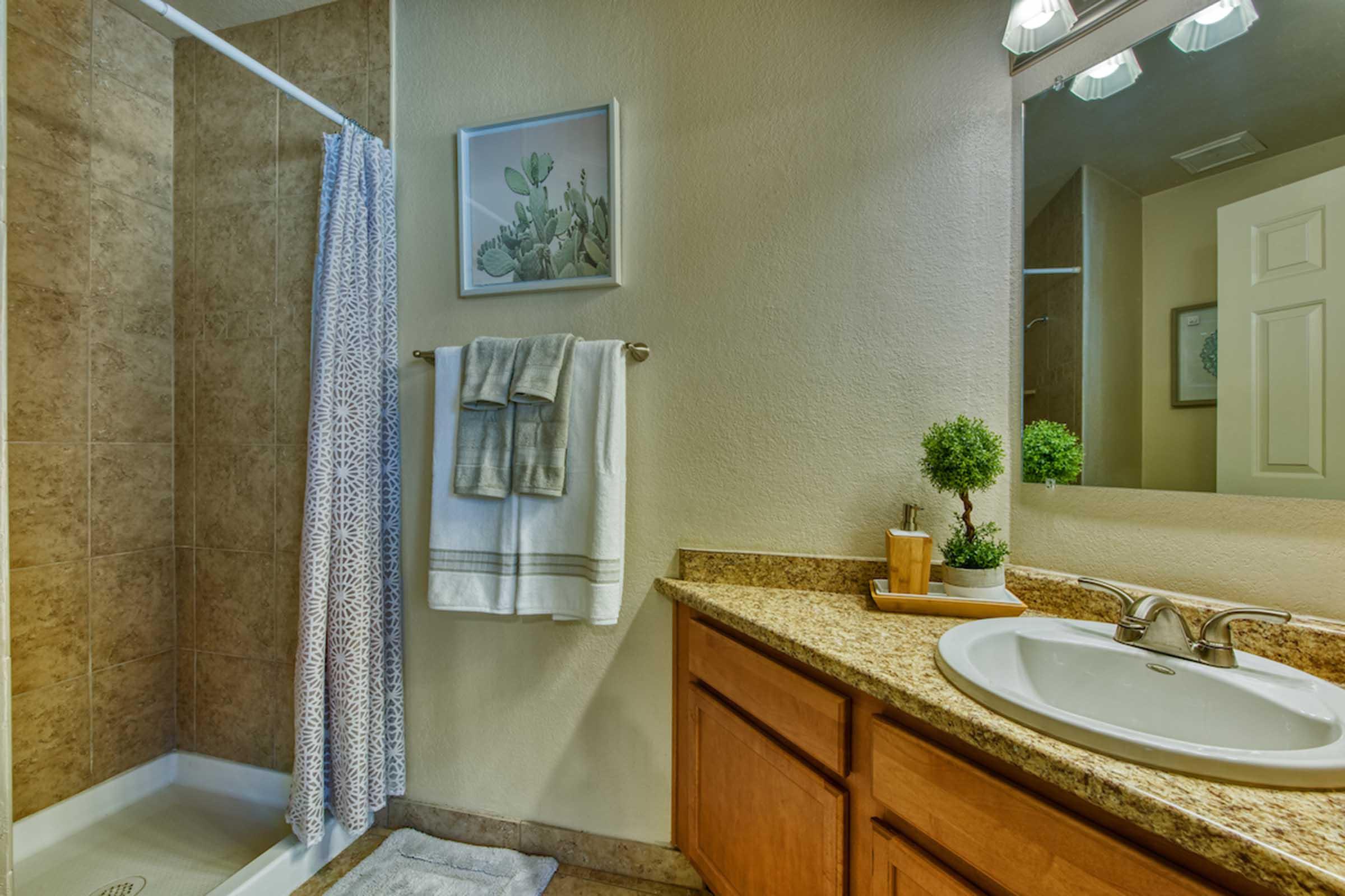 A modern bathroom featuring a shower area with a white curtain, beige tile walls, a granite countertop with a sink, and a large mirror. The decor includes neatly hung towels, a small potted plant, and artwork on the wall, creating a clean and inviting space.