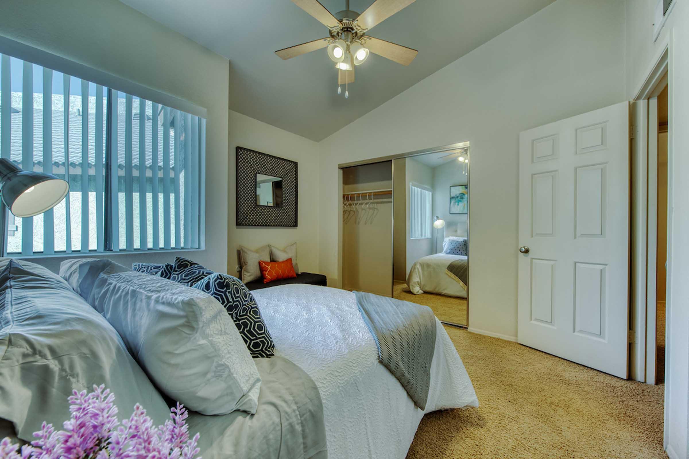 A cozy bedroom featuring a queen-size bed with decorative pillows, a bedside lamp, and a cozy chair in the corner. The room has a large window with vertical blinds, a mirrored closet, and soft carpeting. A small plant adds a touch of color to the space. Natural light brightens the room, enhancing its inviting atmosphere.