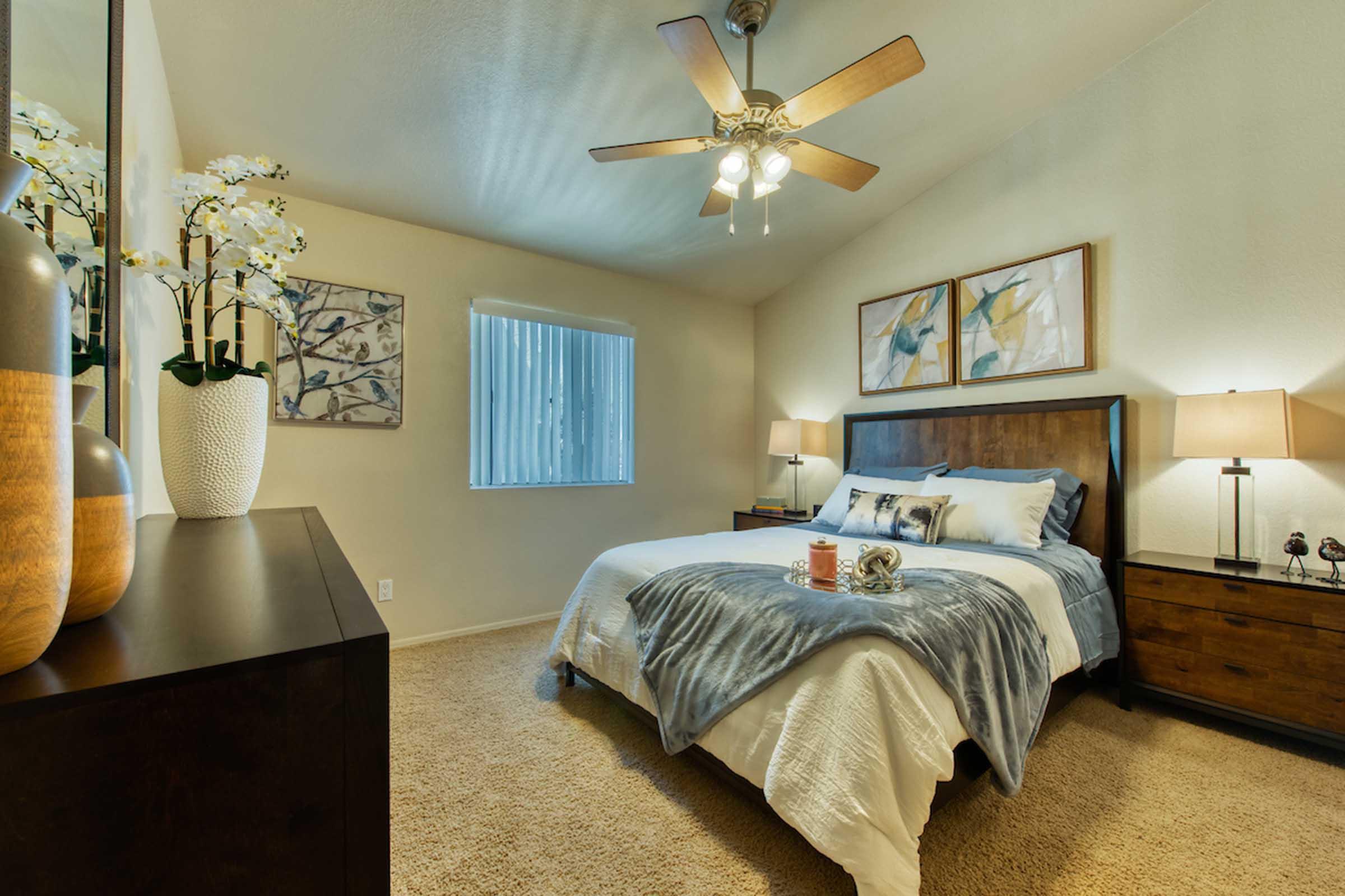 A cozy bedroom featuring a queen-sized bed with decorative pillows, a wooden headboard, and matching nightstands with lamps. A dresser is positioned against the wall, and a window allows natural light to enter, complemented by soft beige walls and a ceiling fan. Decorative flowers add a touch of elegance to the space.
