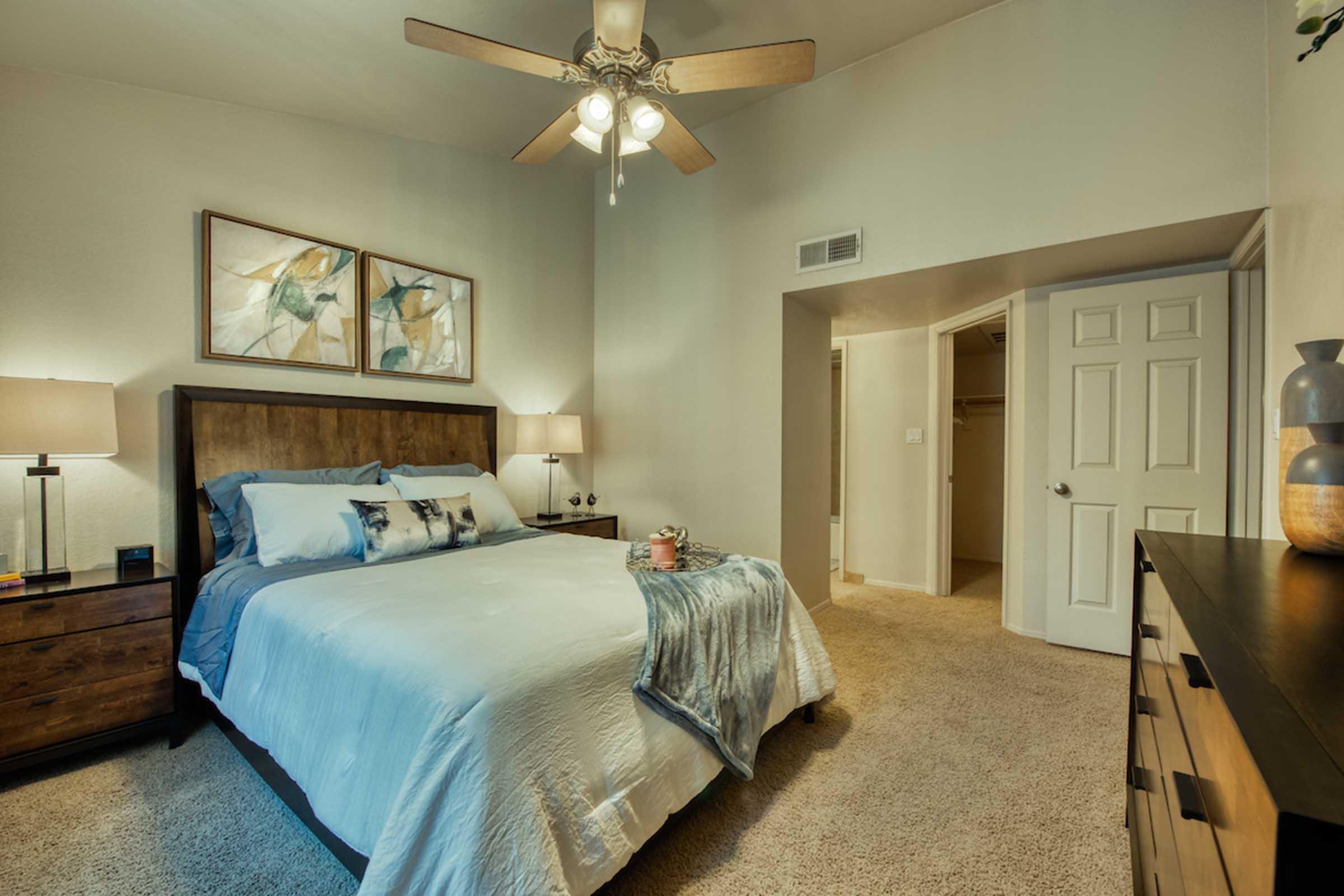 A cozy bedroom featuring a wooden bed frame with a light blue and white bedding set. Two bedside lamps illuminate the space, complemented by framed art on the wall. A ceiling fan hangs above, and a closet is visible through an open door. The room has plush carpet and a warm, inviting atmosphere.