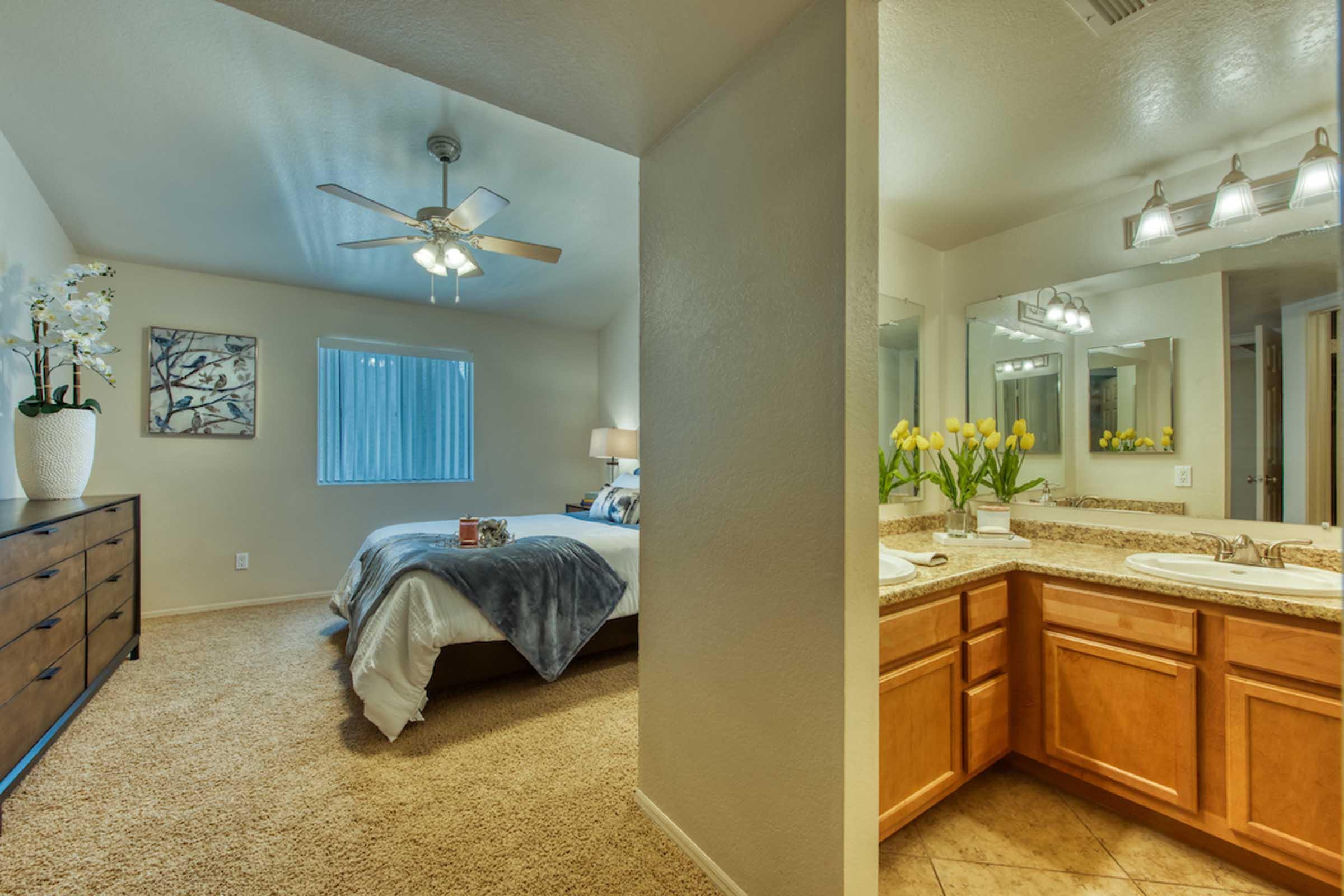 A spacious bedroom with a king-sized bed and light-colored furnishings, visible from an adjoining bathroom. The bedroom features a ceiling fan, a dresser, and a window with blinds. The bathroom has dual sinks, granite countertops, and stylish lighting, with bright yellow tulips adding a decorative touch.