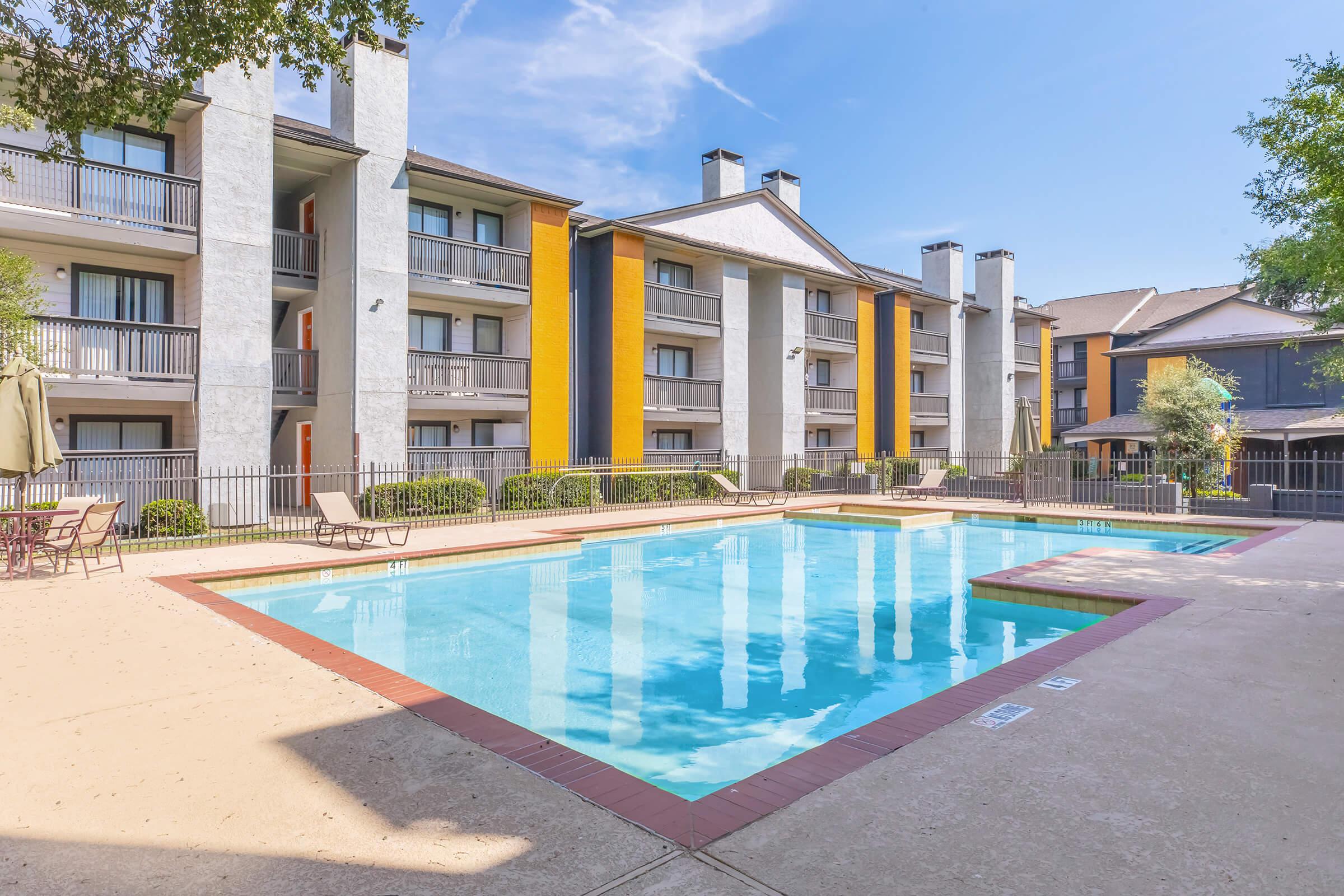 A bright, sunny day at an apartment complex featuring a swimming pool surrounded by chairs and umbrellas. The buildings are three stories tall, with a mix of gray and yellow exteriors. Lush green landscaping enhances the inviting atmosphere. The clear blue sky reflects in the pool water.