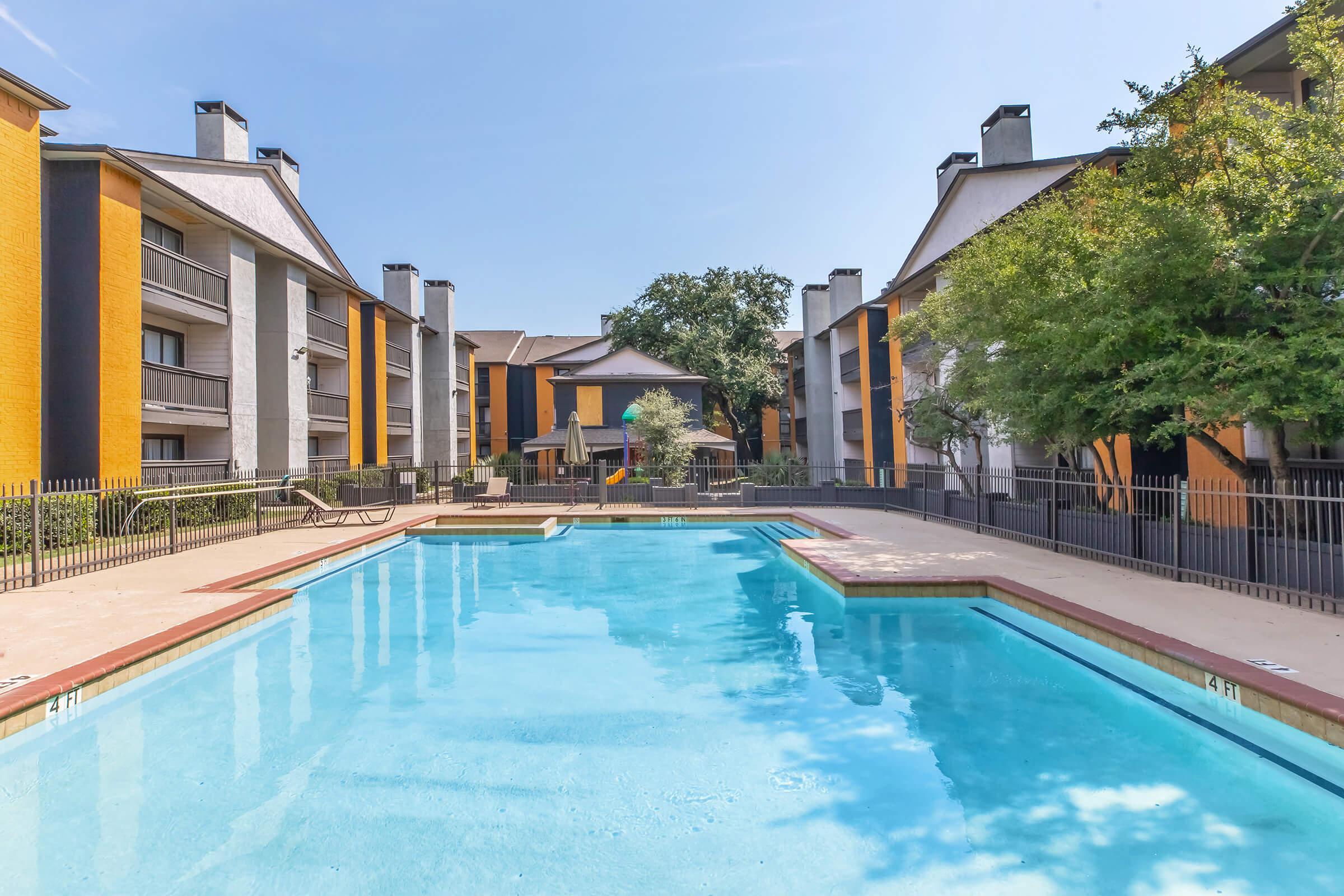 A bright outdoor swimming pool surrounded by residential buildings. The pool has a clean, blue surface and is enclosed by a fence. Nearby, there are lounge areas and greenery, creating a relaxing atmosphere in a well-maintained community setting.