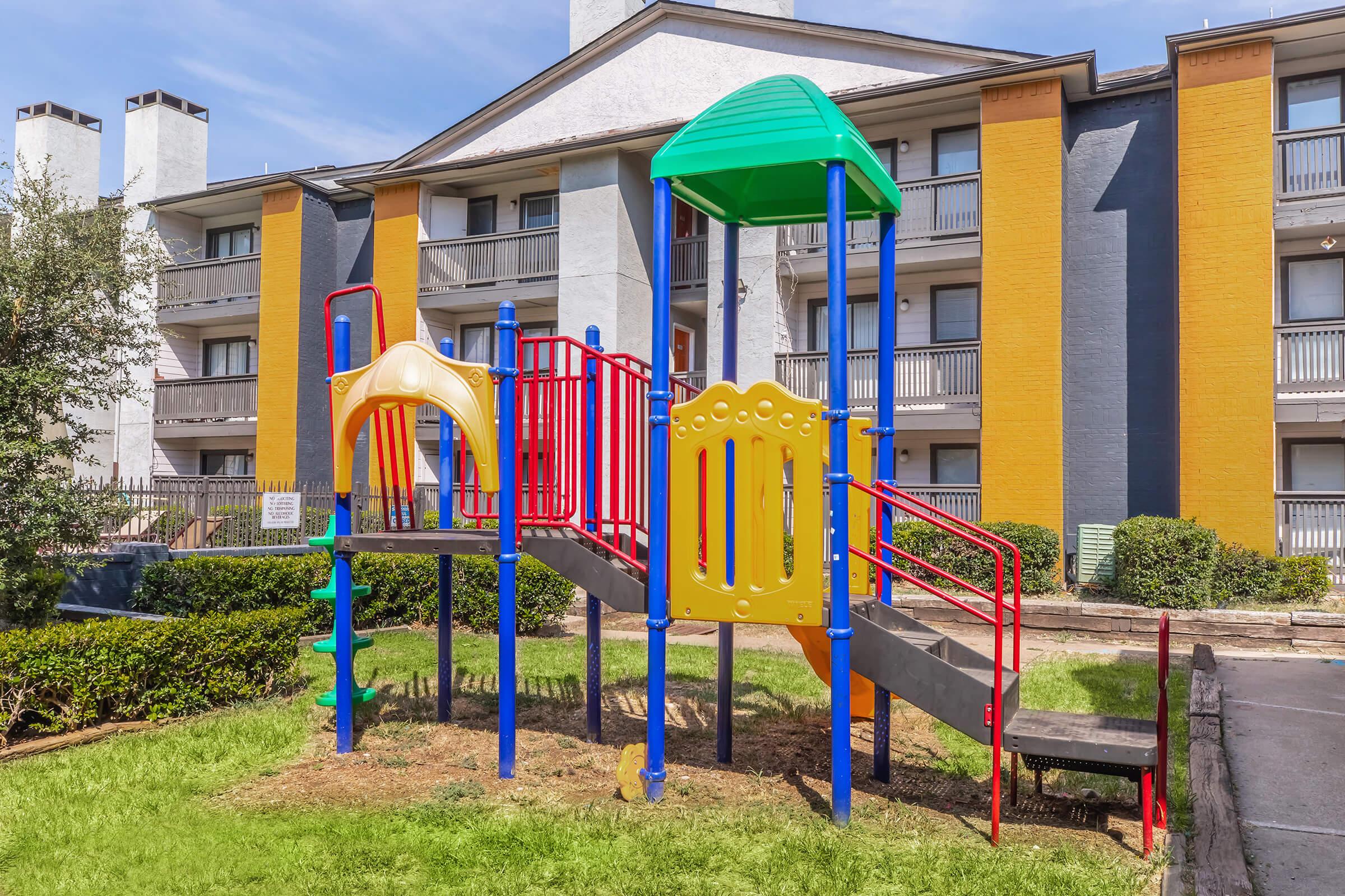 A children's playground featuring a colorful structure with a green roof, yellow slides, and red railings. The playground is situated on a grassy area near a residential building with a modern design, characterized by yellow and gray walls.