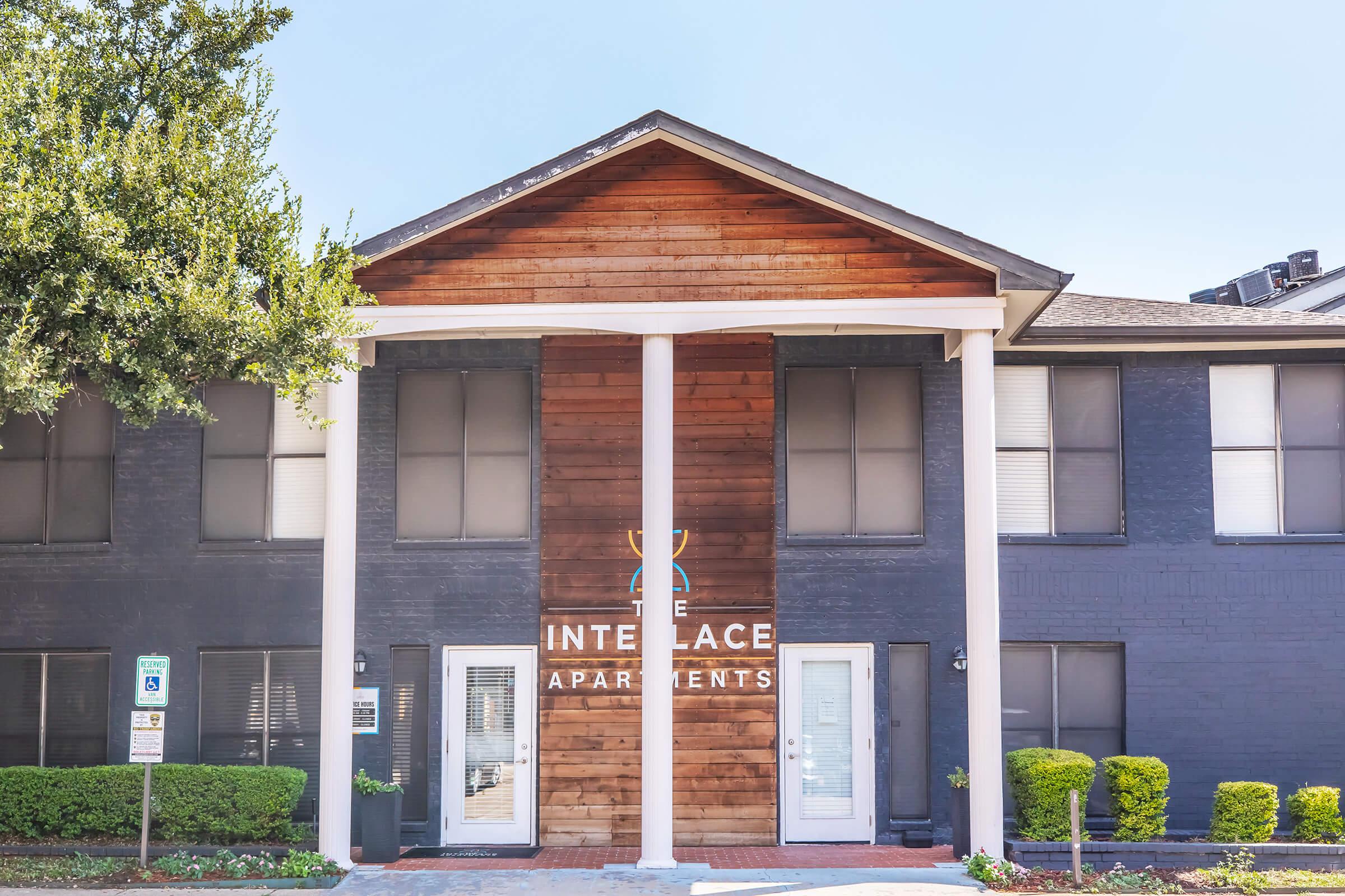 Facade of Intelace Apartments featuring a mix of wooden and brick design, with two entrance doors flanked by large windows. The building has a modern aesthetic, highlighted by a sloped roof and columns supporting the entrance. Lush green bushes border the walkway leading up to the building.