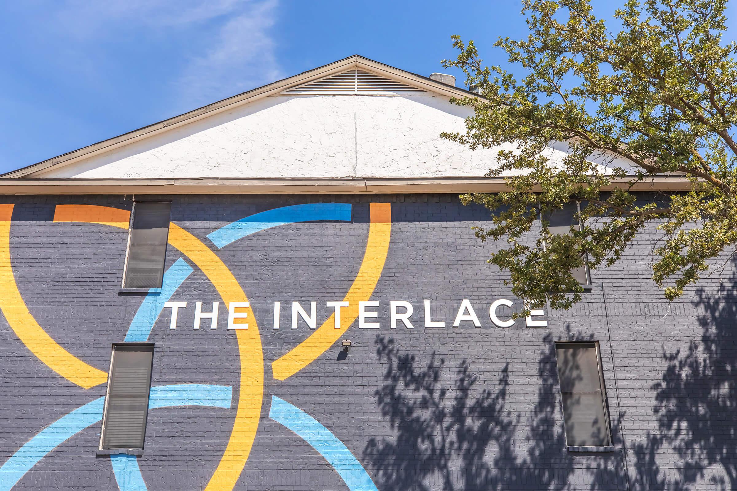 A mural on a building featuring the text "THE INTERLACE" in bold white letters, accompanied by colorful circular designs in yellow and blue. The background is a light gray wall, and a tree is partially visible in the foreground against a clear blue sky.