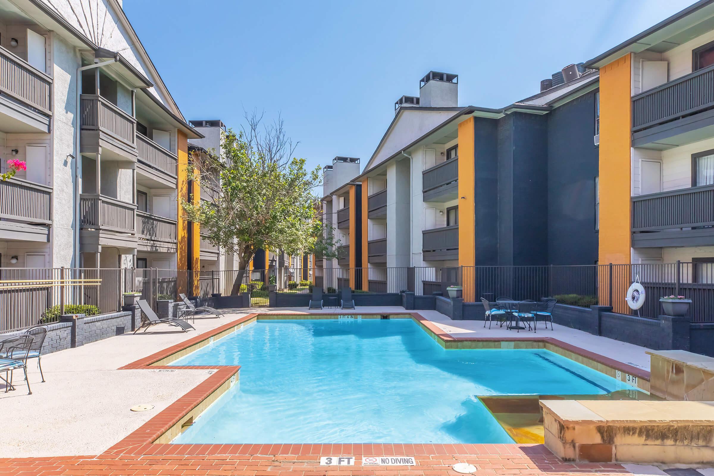 A bright and inviting apartment complex featuring a central swimming pool surrounded by lounge chairs. The buildings are two stories tall, with a mix of light and dark colors. Trees are visible, providing some shade, and the sky is clear and sunny, contributing to a relaxed outdoor atmosphere.