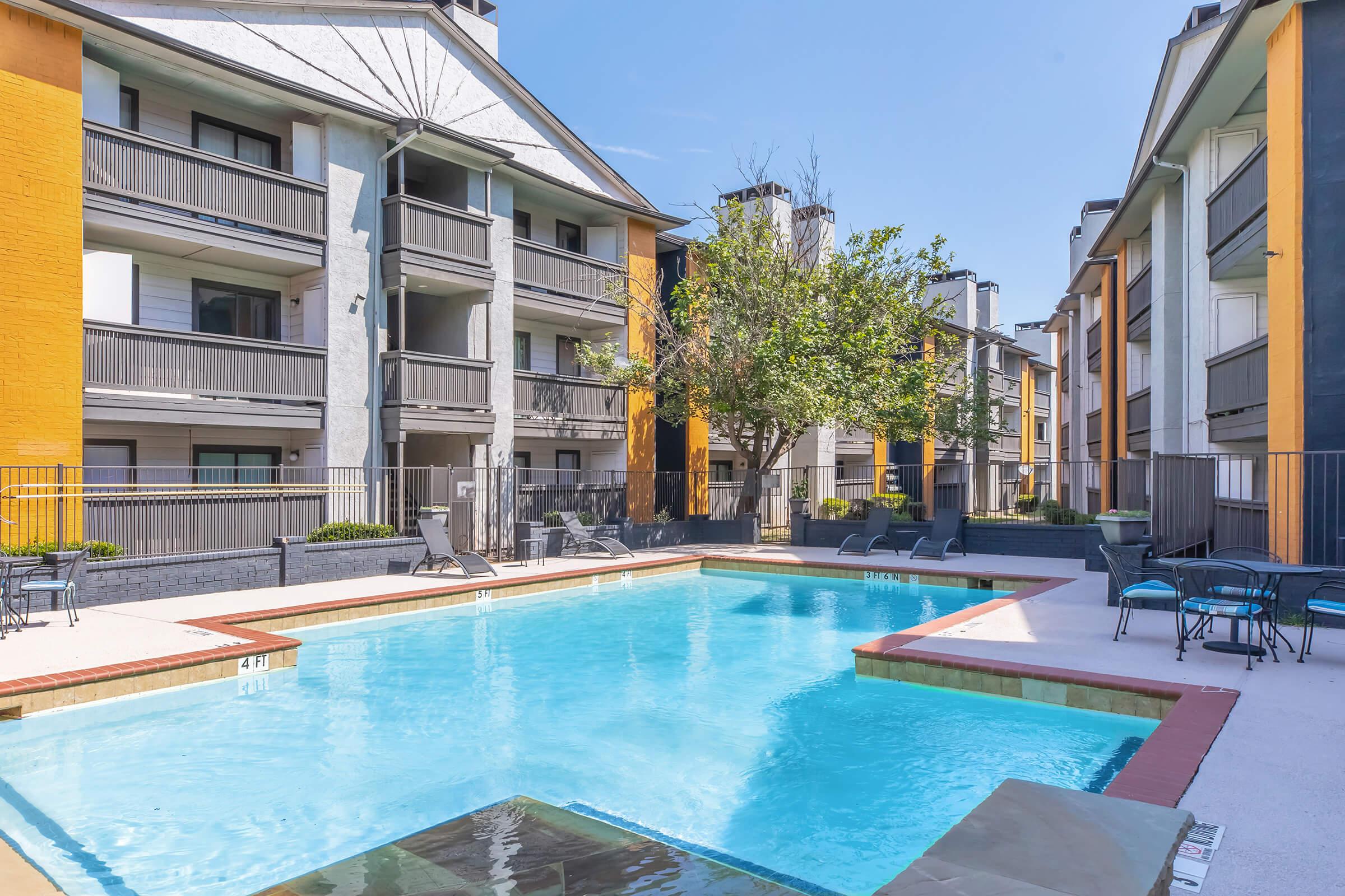 A well-maintained apartment complex features a sparkling blue swimming pool surrounded by lounge chairs, with buildings displaying modern architecture in the background. Lush greenery and clear blue skies add to the inviting atmosphere.