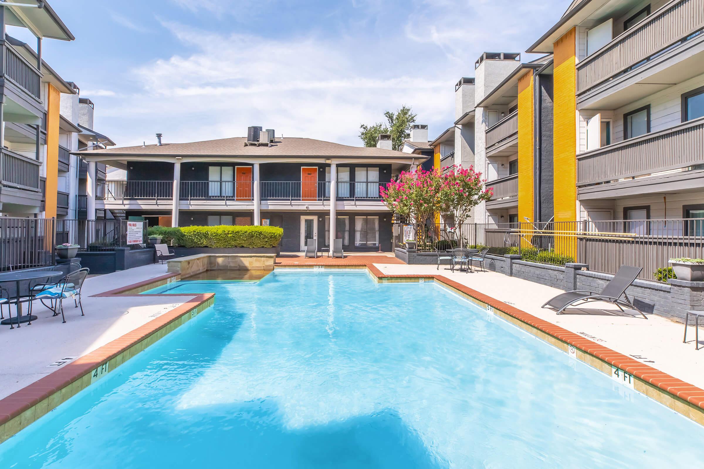 A clear blue swimming pool surrounded by a patio with lounge chairs, situated in the courtyard of a modern apartment complex. The building features multiple levels with balconies, and vibrant landscaping with flowering plants adds a touch of color. The sky is bright with a few clouds.