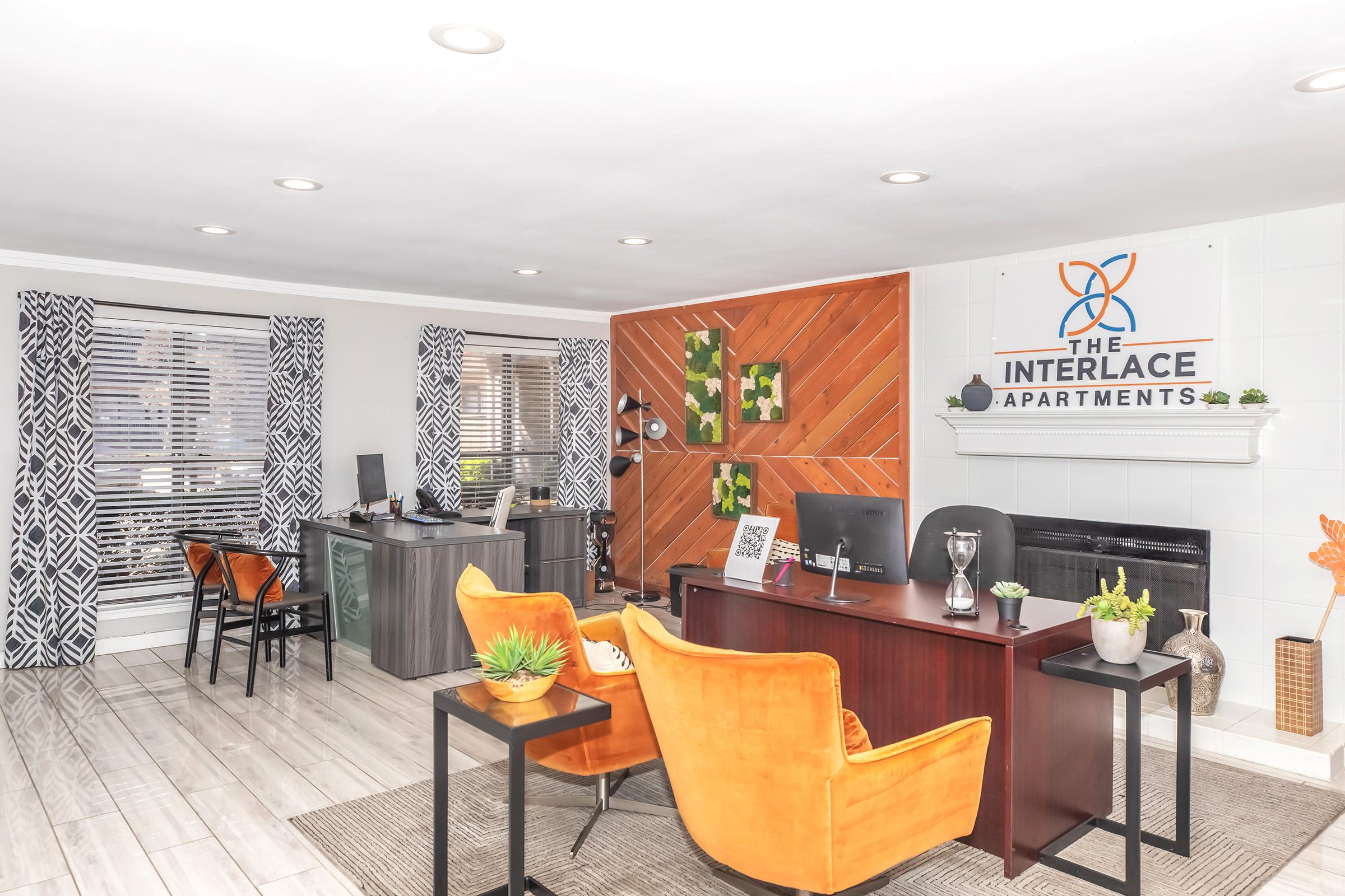 Interior of an apartment lobby featuring a reception area with dark wood furniture, orange chairs, and decorative plants. The walls have patterned curtains and artwork. A sign reads "THE INTERLACE APARTMENTS." The floor is light-colored tile, creating a welcoming atmosphere for residents and visitors.