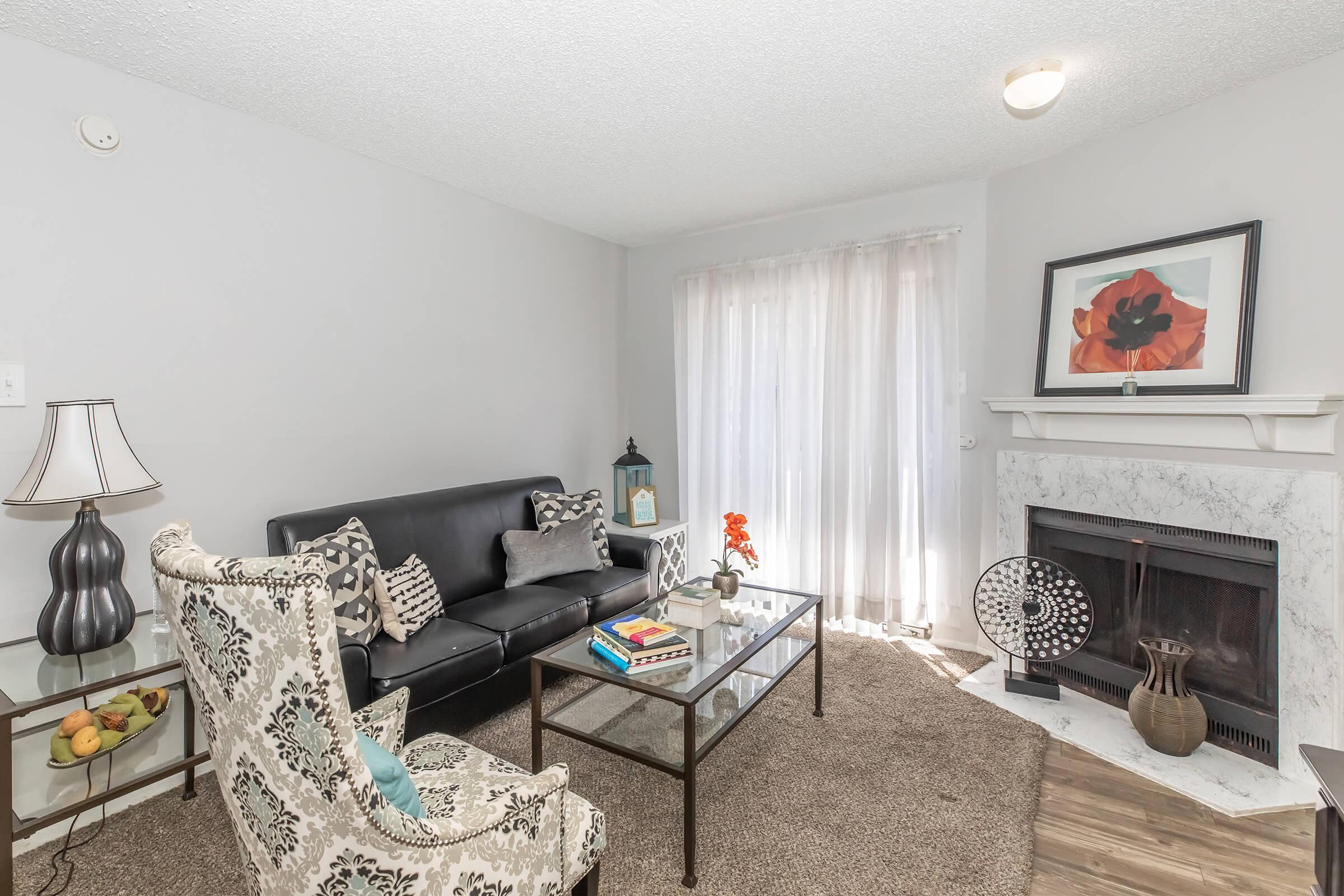 A cozy living room featuring a black leather sofa, a decorative armchair with a floral pattern, and a glass coffee table holding books and a small vase with flowers. A fireplace with a white mantel is visible, and large windows allow natural light to enter, complemented by sheer curtains.