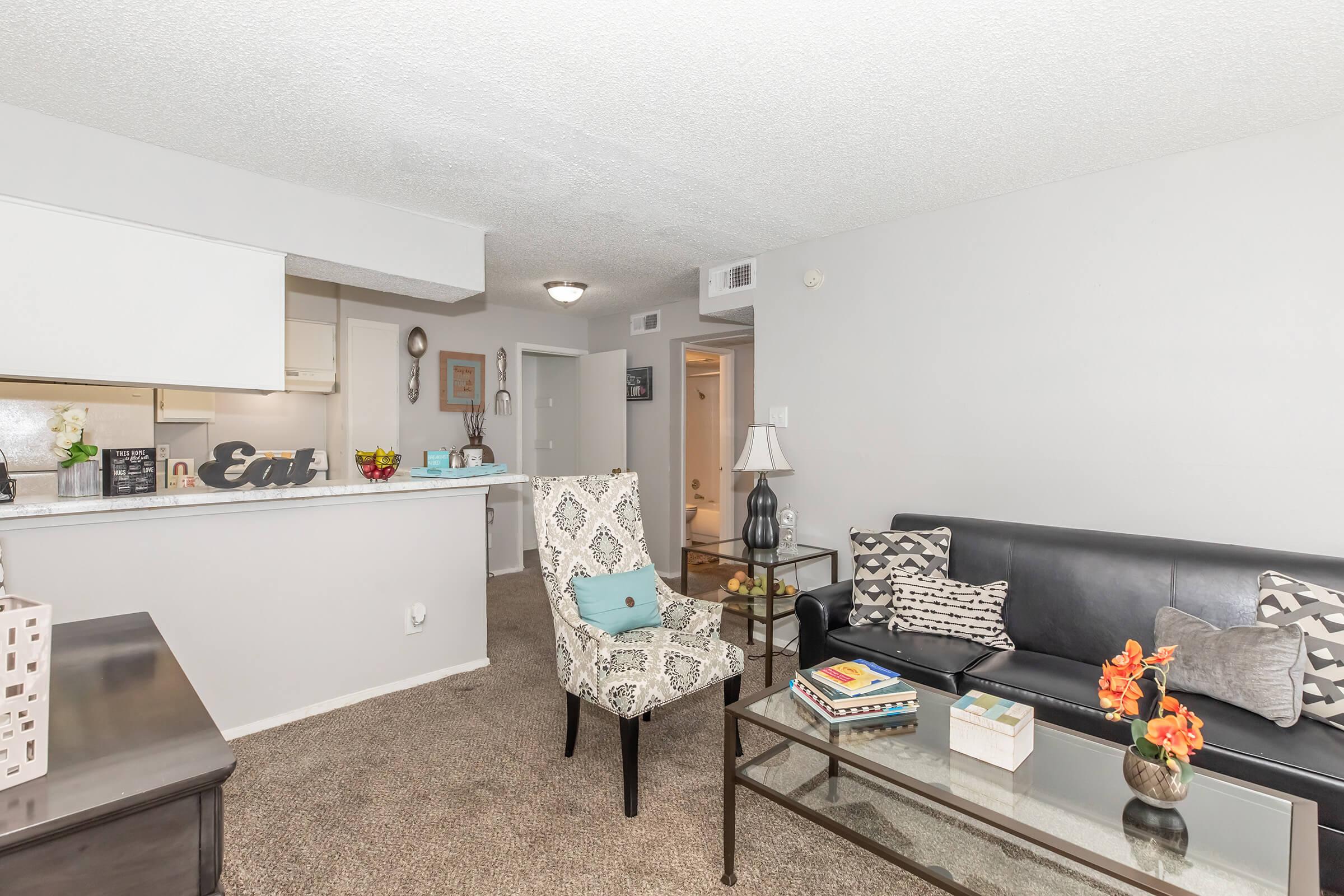 Cozy living room featuring a plush black sofa and a stylish patterned armchair. A glass coffee table sits on a soft carpet, adorned with decorative items and flowers. The open layout includes a kitchen area in the background, creating a warm and inviting atmosphere.