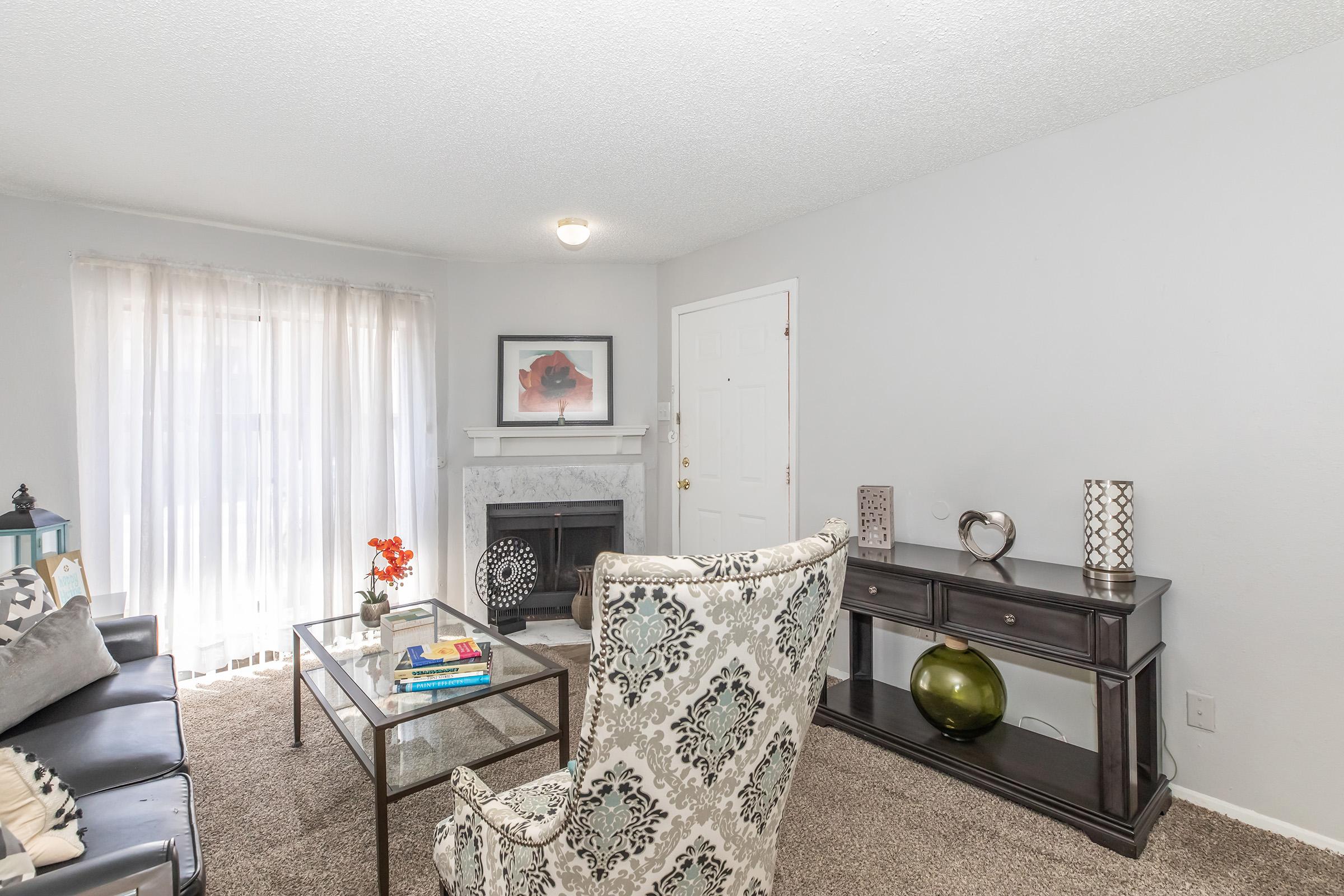 Cozy living room featuring a gray couch with decorative pillows, an elegant armchair, a glass coffee table with books and decor, a fireplace, and a console table. Natural light flows through sheer curtains, creating a welcoming atmosphere.