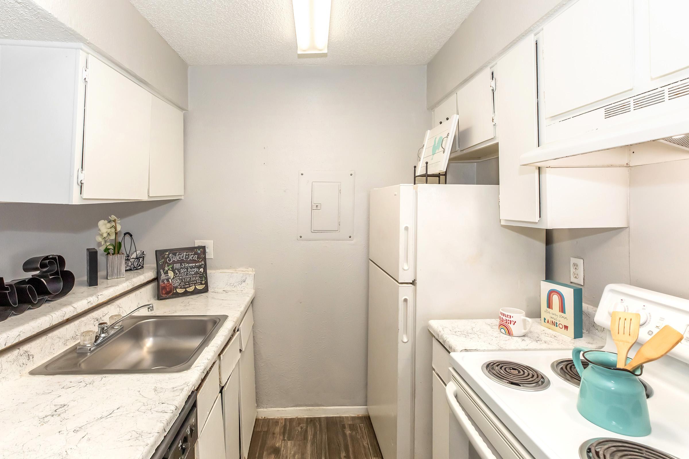 A small kitchen featuring white cabinets, a stainless steel sink, a white refrigerator, and a stove. The countertops are light-colored with decorative items, including a chalkboard sign and a teal kettle. The walls are painted gray, and there is a window above the sink providing natural light.