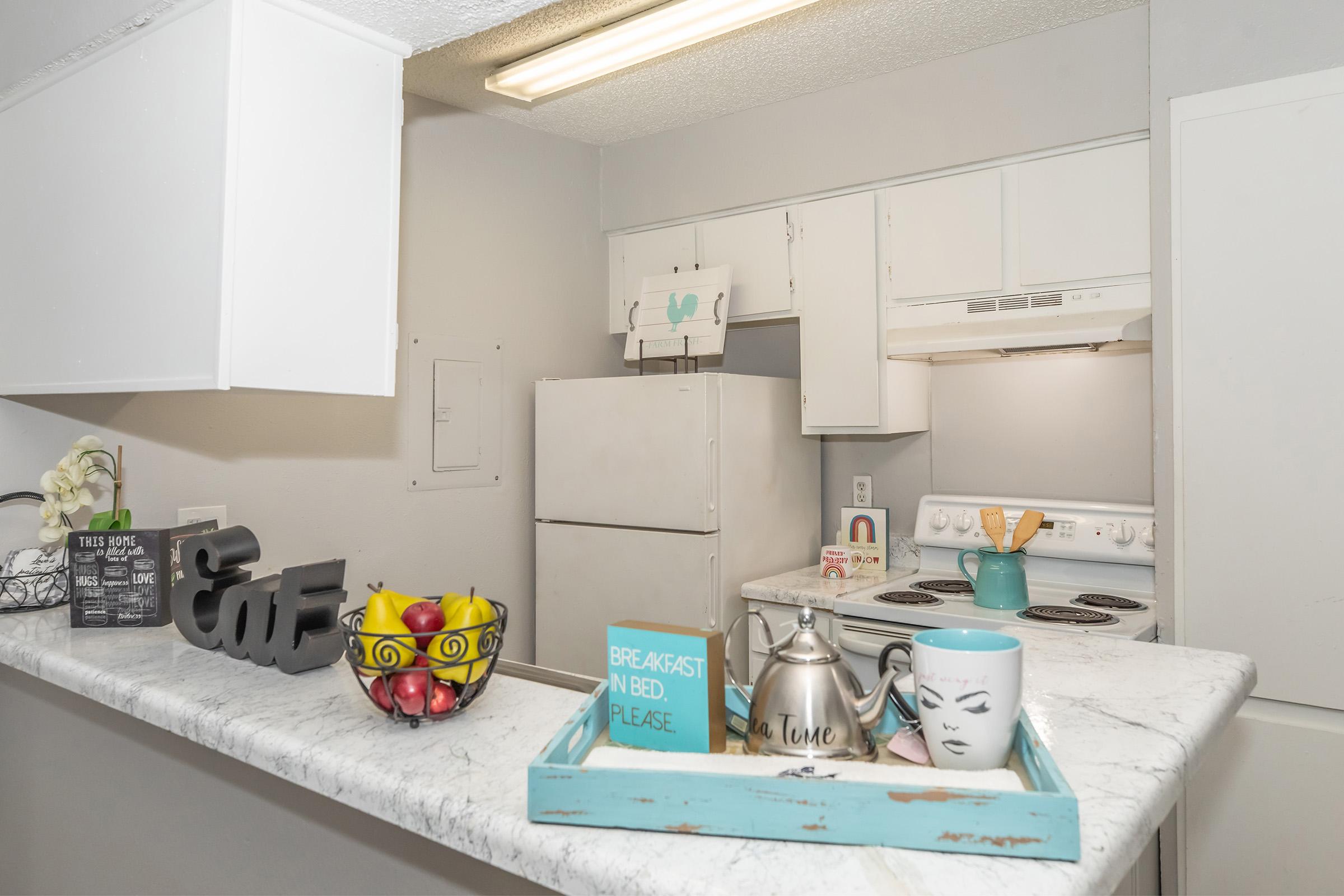 A modern kitchen with light gray walls and a marble countertop. There is a white refrigerator, stove, and various kitchen items. A decorative tray holds a teal mug with a face design, a teapot, and fresh fruit. A small sign reads "BREAKFAST IN BED, PLEASE," adding a cozy touch to the space.