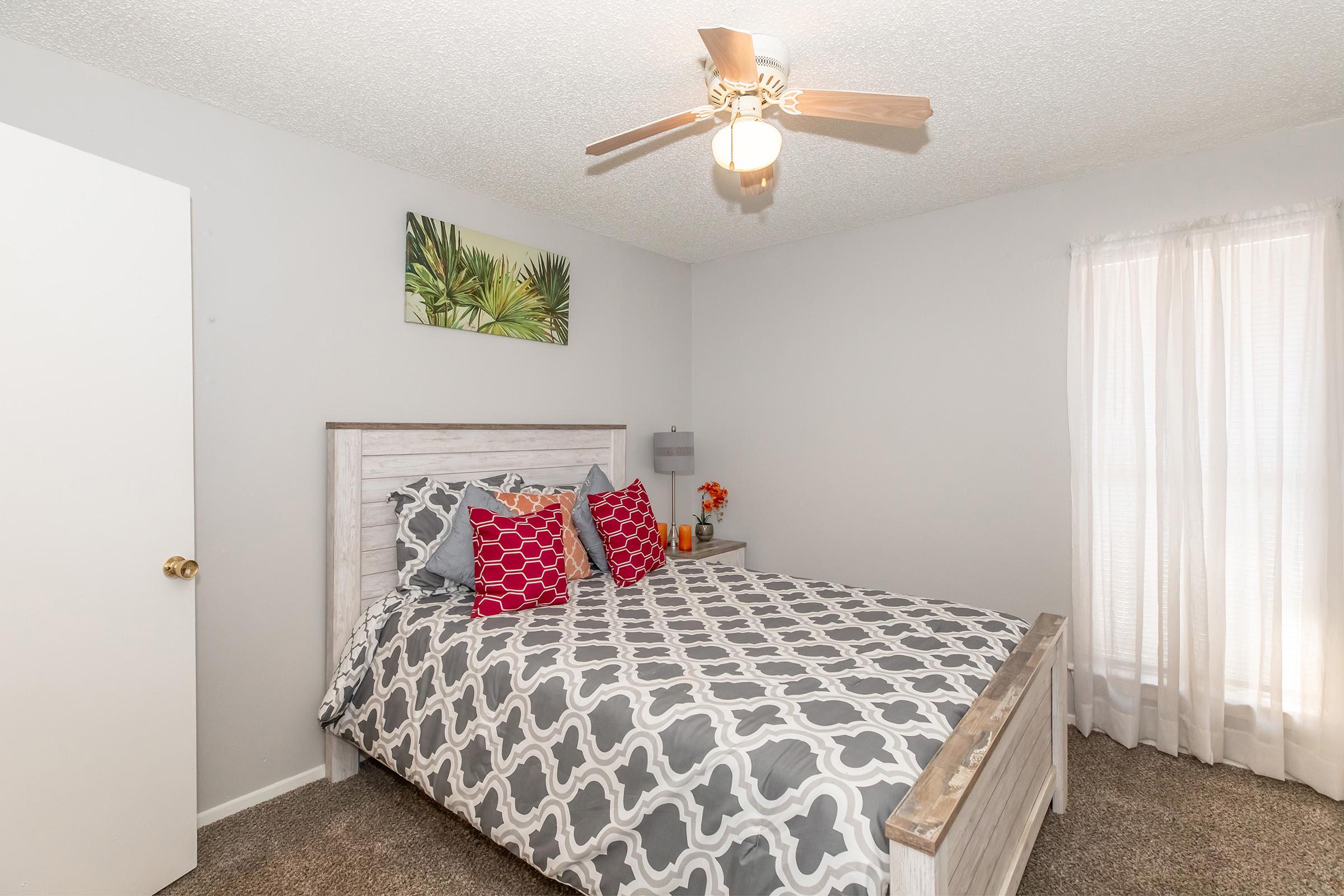 A cozy bedroom featuring a queen-sized bed with patterned gray and white bedding, decorative red and white pillows, a ceiling fan, and a window with sheer white curtains. A piece of tropical wall art adds a pop of color, and a small nightstand with a lamp and a vase of flowers completes the ambiance.