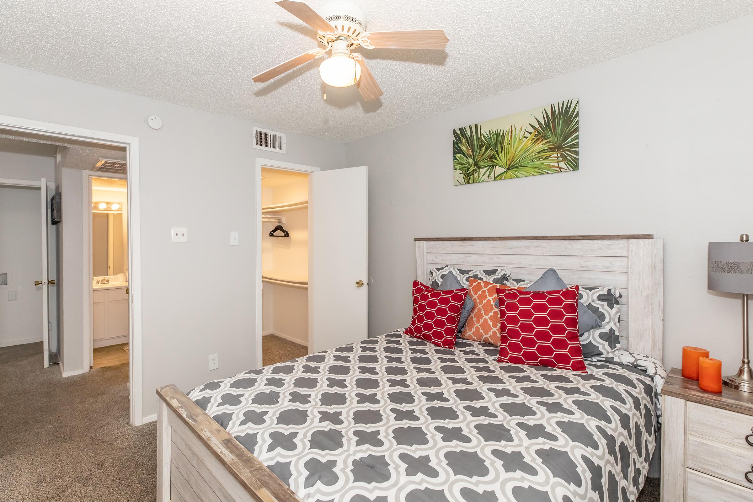 A cozy bedroom featuring a queen-sized bed with a decorative gray and white patterned comforter, colorful accent pillows, and a stylish wooden headboard. A ceiling fan hangs above, and there is a lamp on a nightstand. The room has light gray walls and includes a closet space, with a bathroom visible in the background.