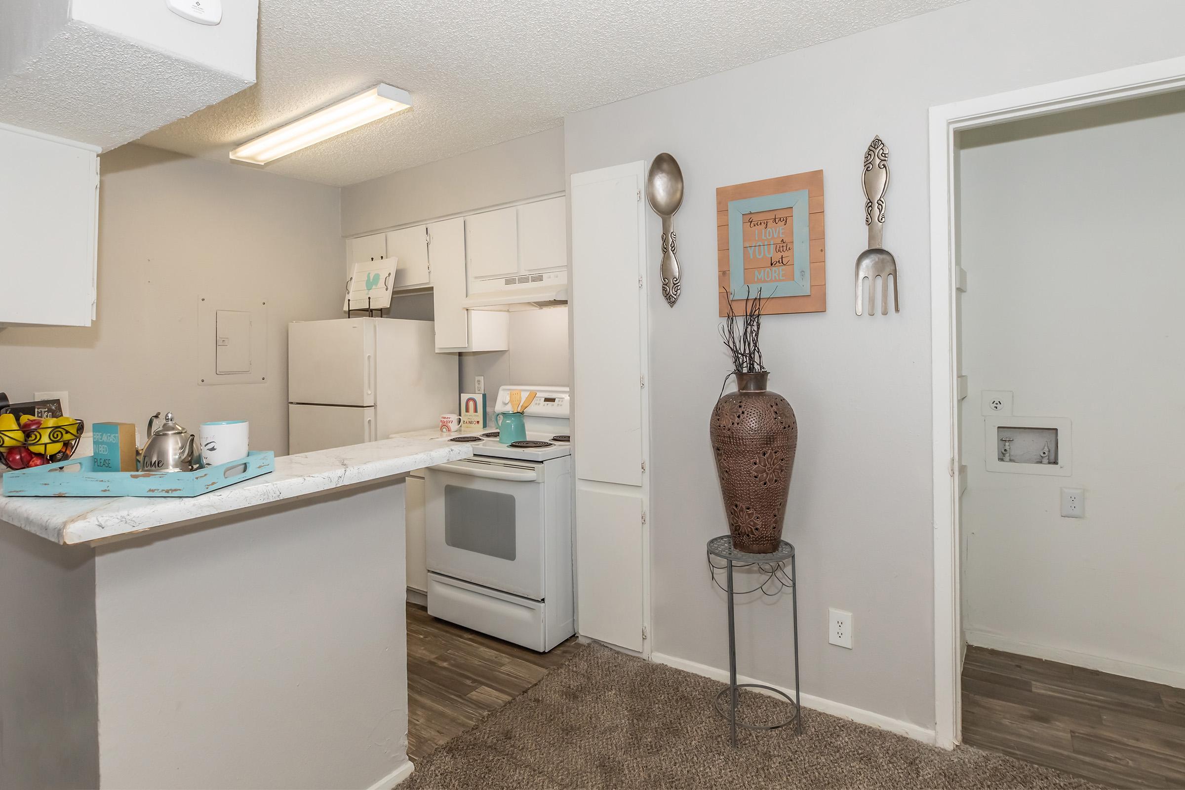 A modern kitchen featuring white cabinetry and appliances, a countertop with a small teapot, and decorative items. There's a textured brown vase with branches and wall art with kitchen-themed decor. The flooring is wood-like, and the space is well-lit with overhead lighting.
