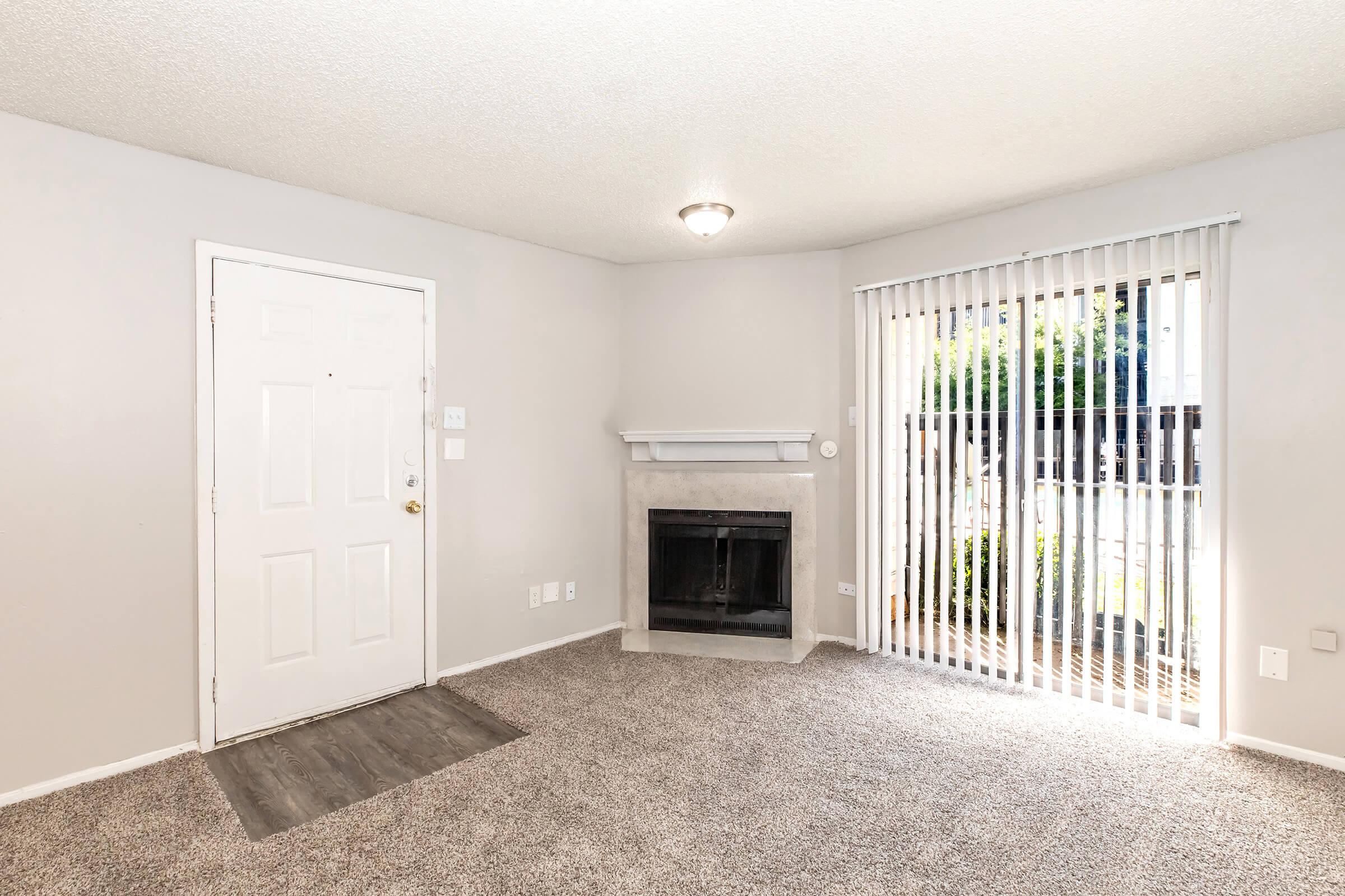 A cozy living room featuring a light gray wall and carpet, a front door on the left, and a fireplace with a mantel. There are vertical blinds covering a large window that lets in natural light, leading to an outdoor space. The room has a warm and inviting atmosphere.