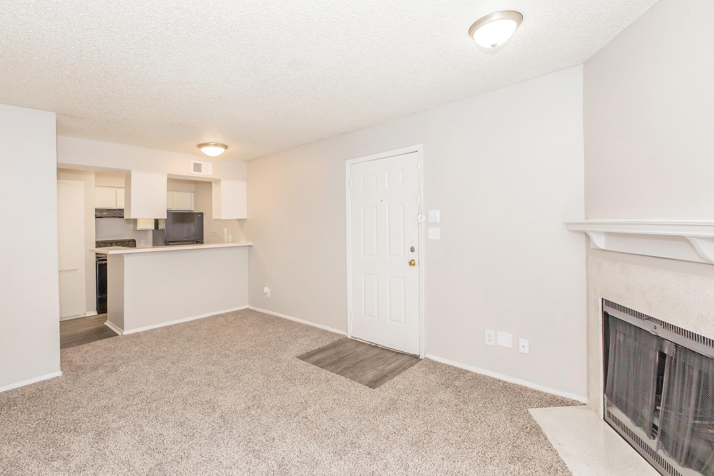 A spacious living area with light beige carpet and neutral-colored walls. The room features a fireplace with a white mantel and a front door. In the background, a kitchen area is partially visible with a countertop and appliances, creating an open and inviting layout.