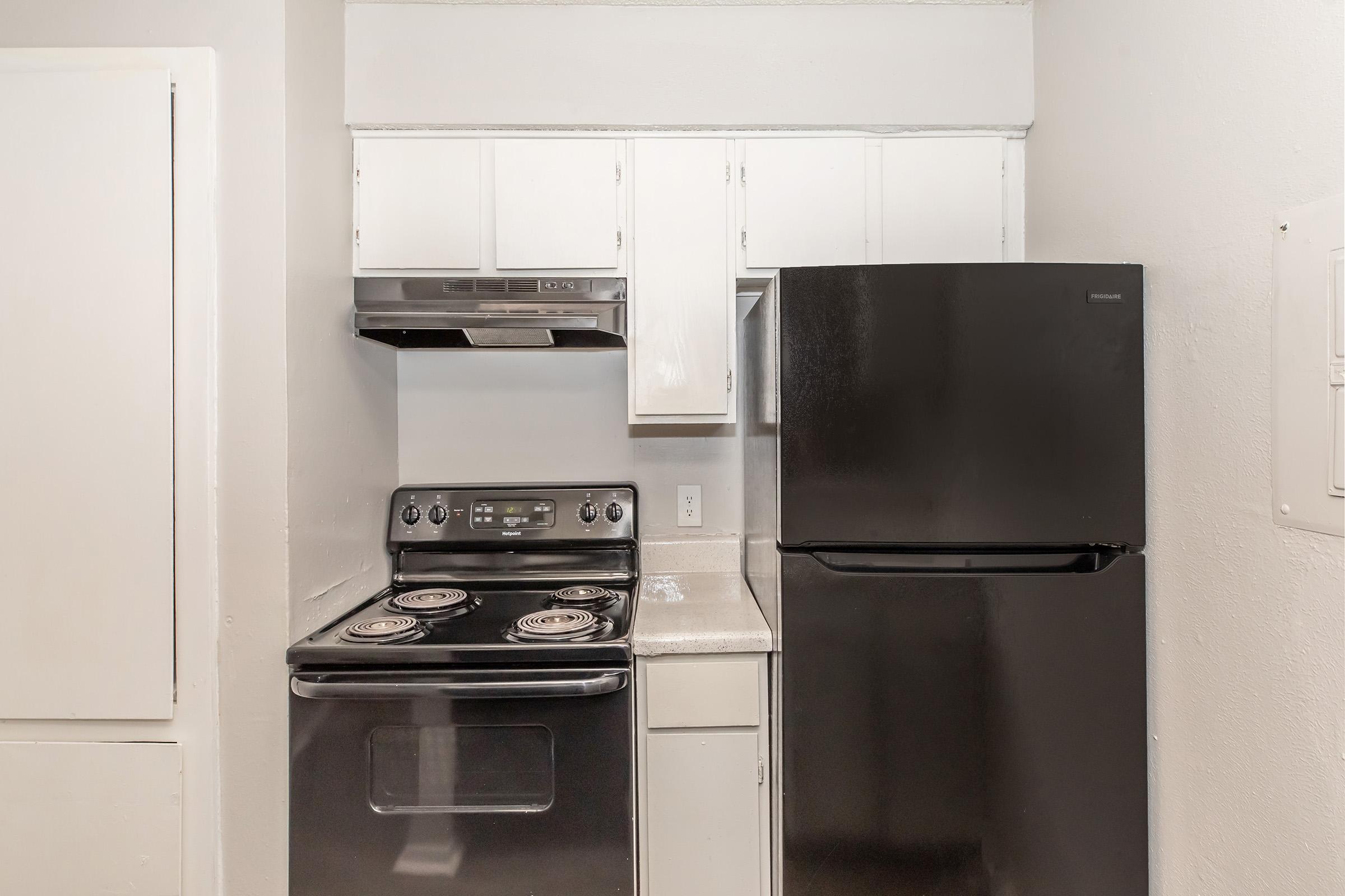 A small kitchen featuring a black refrigerator, an electric stove with four burners, and a hood above. There are white cabinets above the countertops, and the overall design is simple and compact. The walls are painted light colors, contributing to a clean, modern look.