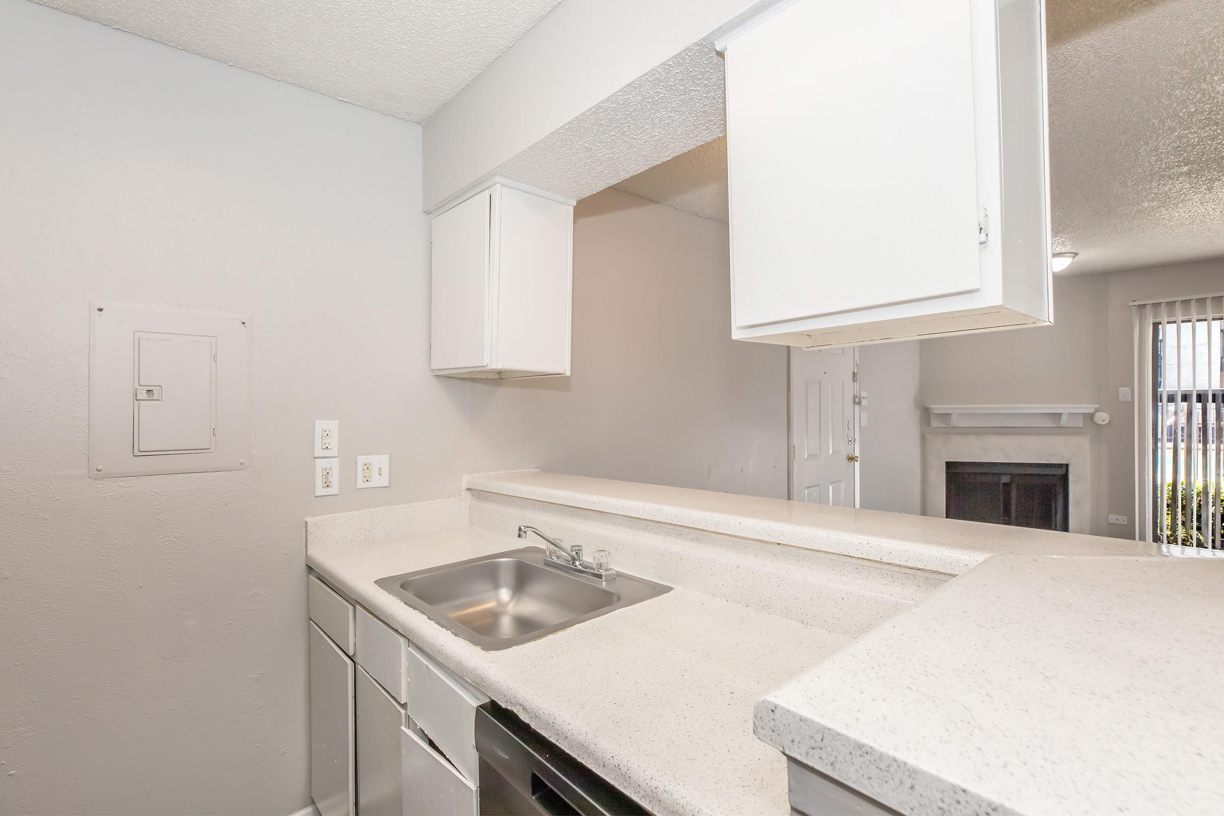 A clean and modern kitchen with a small sink, white cabinets, and a light-colored countertop. The open layout includes a view of a living space with a fireplace and sliding doors that provide natural light. The walls are painted in a soft gray color, creating a neutral and inviting atmosphere.
