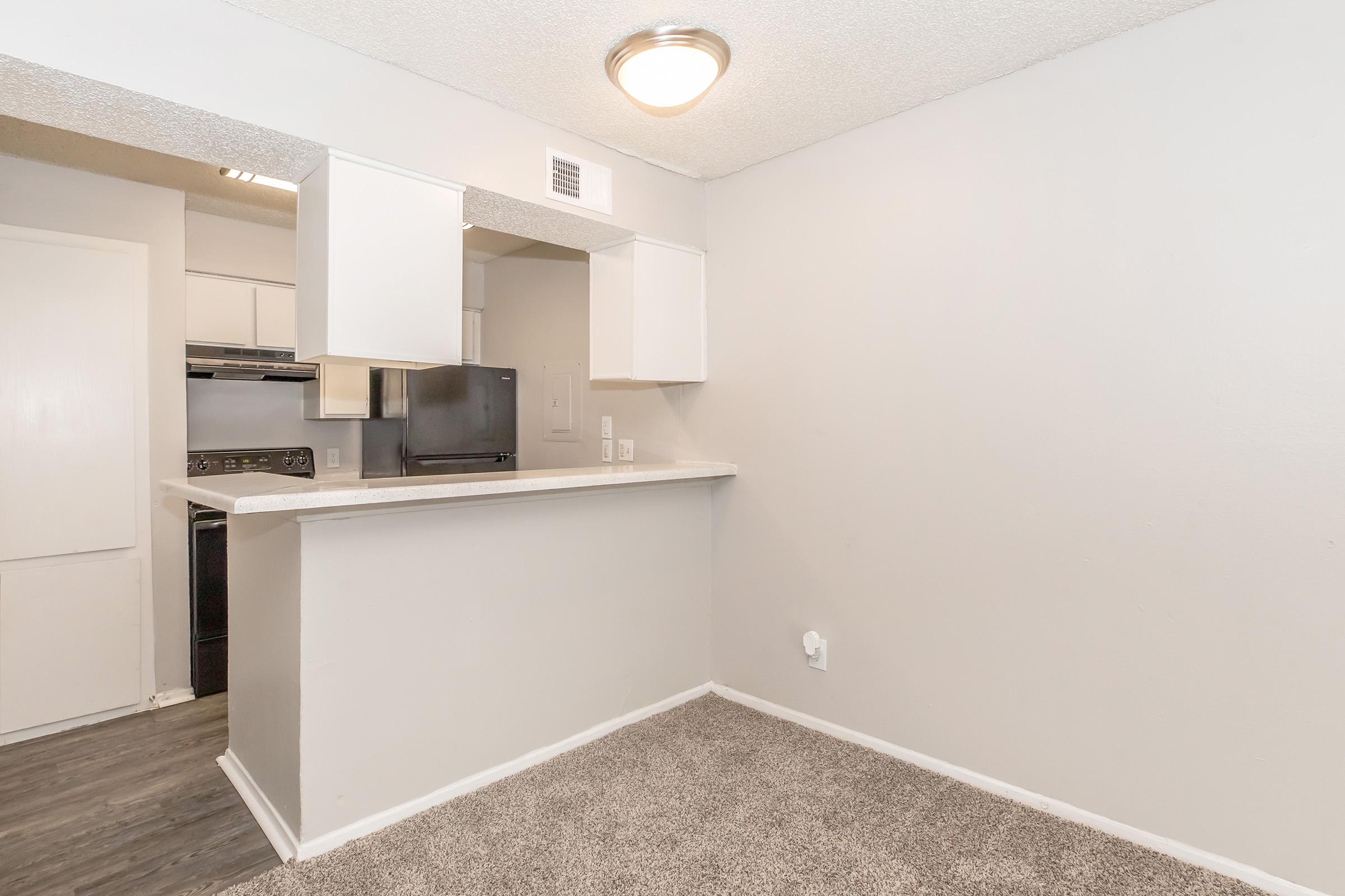 A small, empty living space featuring a light grey wall. On one side, there's a kitchen area with white cabinets, a black refrigerator, and a stove visible. A countertop separates the kitchen from the living area, which has carpeted flooring. The overall ambiance is clean and modern, with natural light from the ceiling fixture.