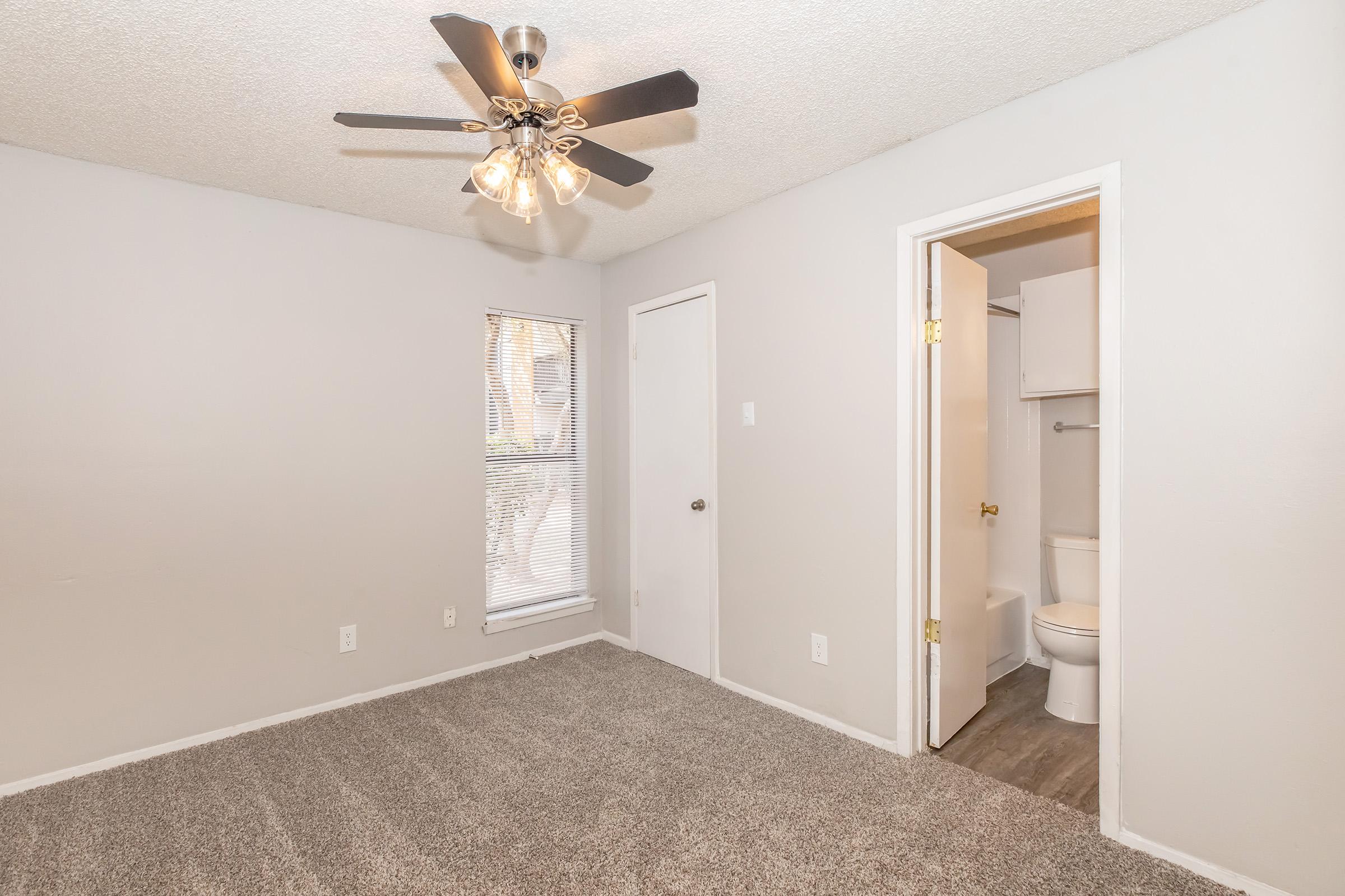 A well-lit bedroom featuring a ceiling fan with light fixtures, gray walls, and plush carpet. There is a window with blinds allowing natural light, and an open door leading to a bathroom area, visible through the doorway. The overall space is clean and inviting, ideal for relaxation.