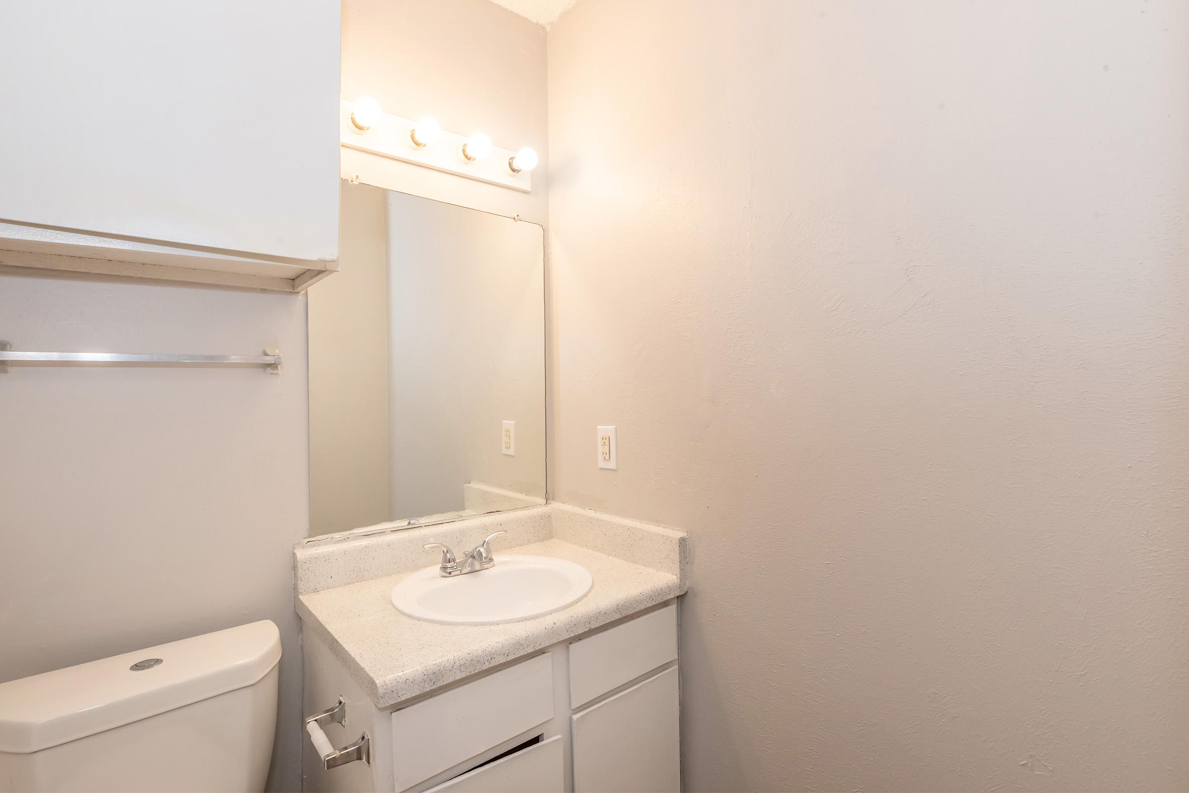 A small bathroom featuring a white sink with a chrome faucet, a large mirror above it, a light fixture with five bulbs, a gray wall, and a white toilet. No decorative elements are visible, creating a clean and minimalist appearance.