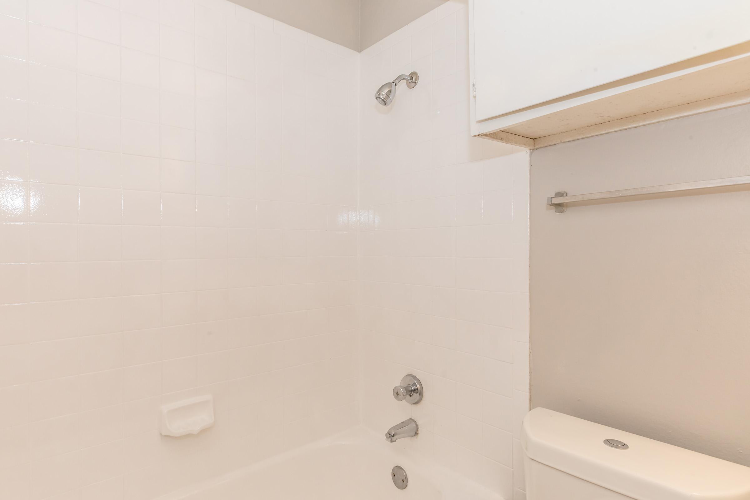 A clean and simple bathroom featuring a white tiled shower area with a showerhead and faucet controls. There is a white bathtub, toilet, and a small overhead cabinet above the bathtub. The walls are painted in a light gray color, contributing to a minimalist aesthetic.