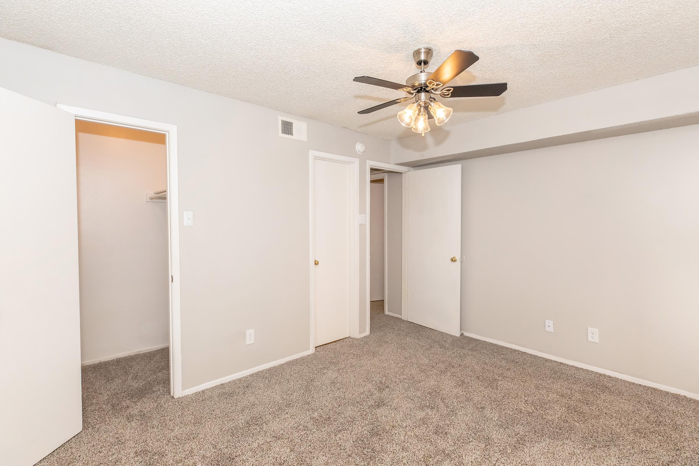A spacious room with light gray walls and carpeted flooring. It features a ceiling fan with four light bulbs and two white doors leading to closets. One door is open, revealing a small closet space. The room is well-lit and tidy, suitable for various uses, such as a bedroom or office.