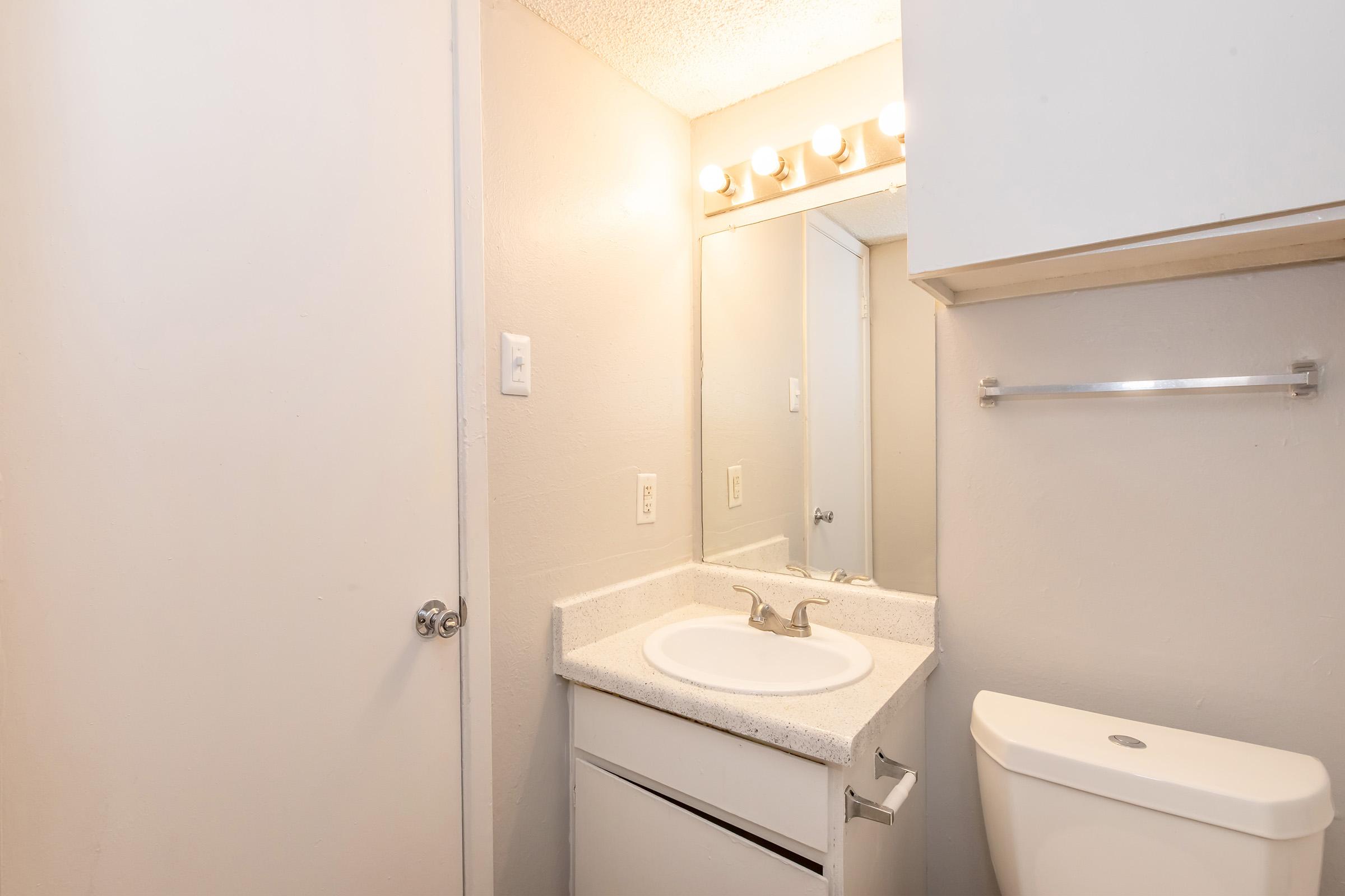 A small bathroom featuring a white sink with a countertop, a mirrored cabinet above the sink, a light fixture with multiple bulbs, a toilet, and a door on the left. The walls are painted light gray, and there is a towel rack next to the sink.