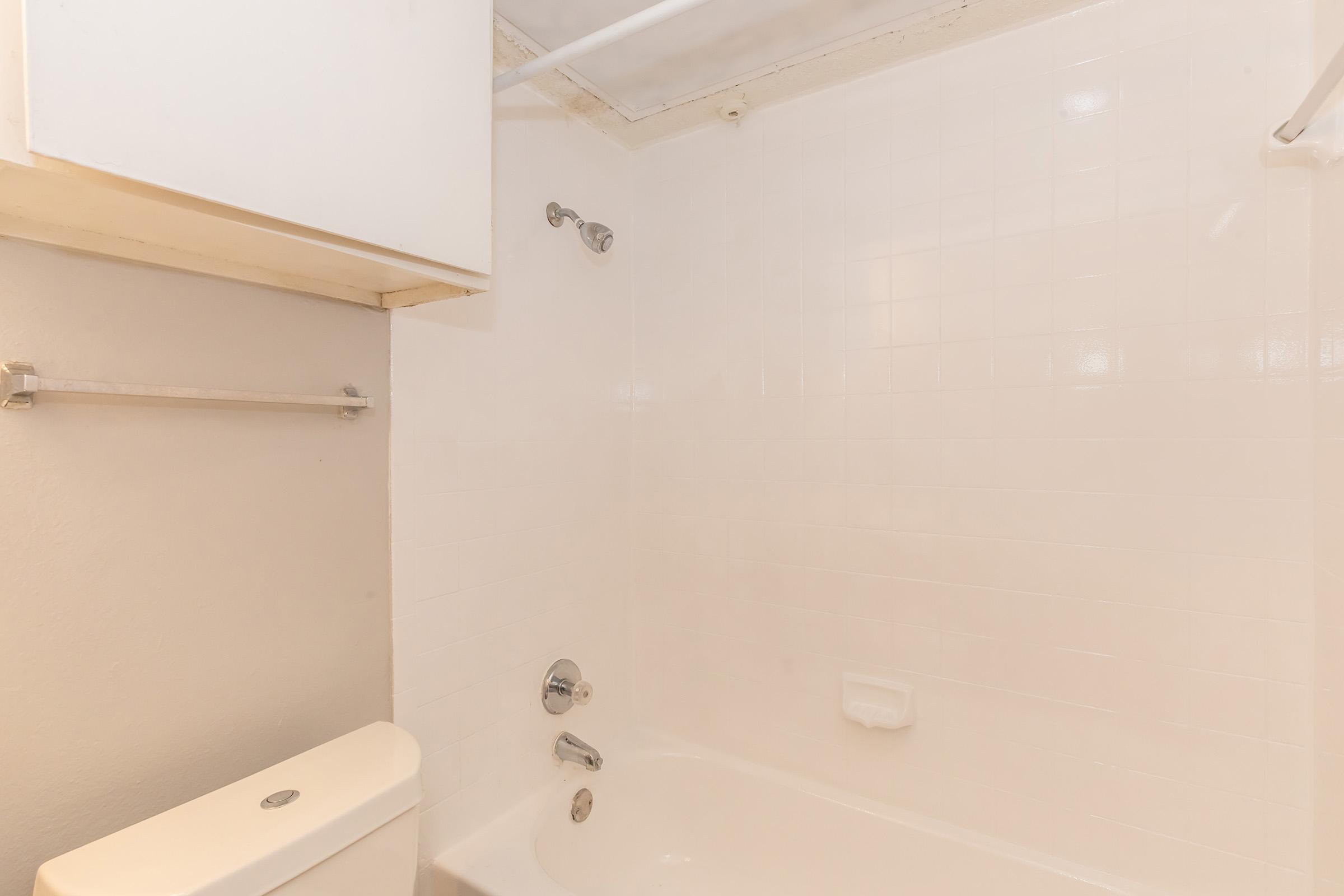 A clean, simple bathroom featuring a white tub, a showerhead, a toilet, and wall-mounted storage. The walls and tiles are also white, creating a bright and spacious look. There's a towel rack and a small shelf above the toilet.